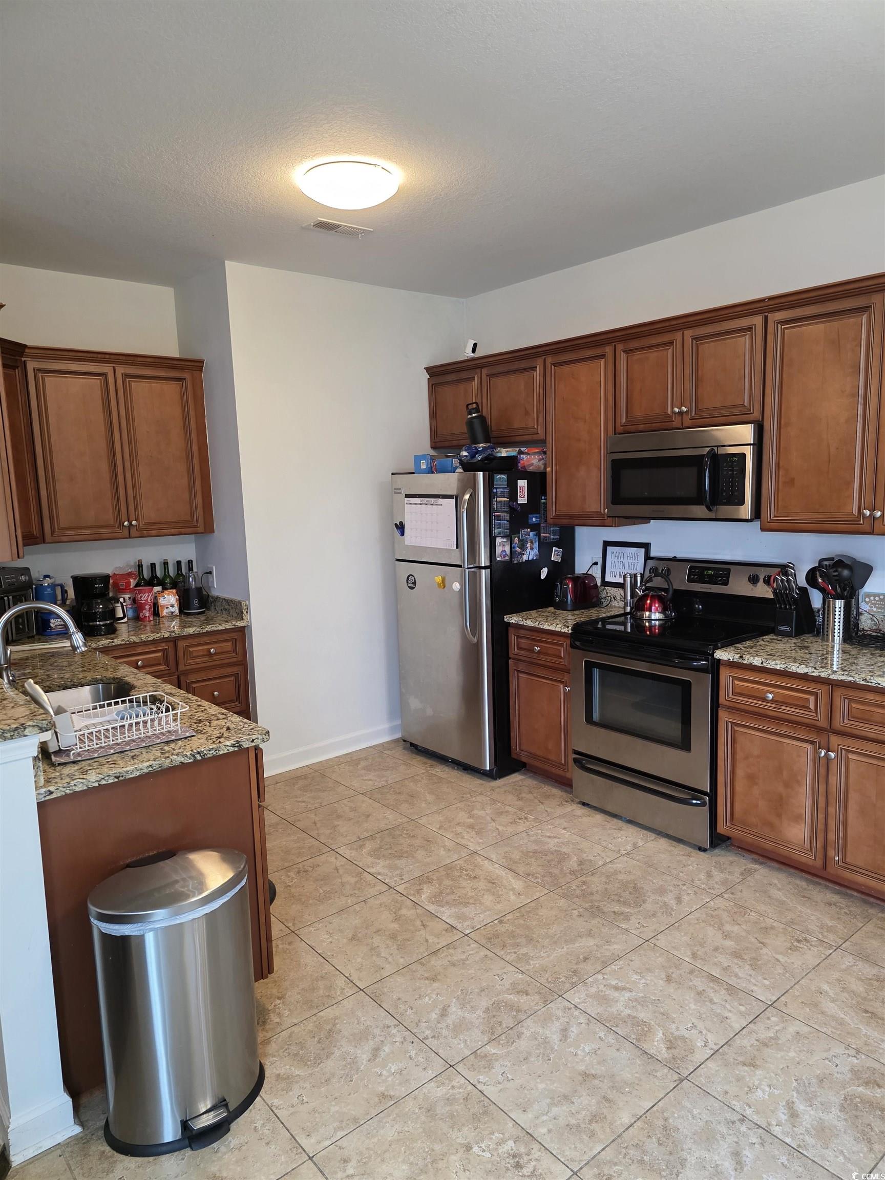 417 Cassian Way Myrtle Beach, SC 29588 - Photo 9 of 13 Kitchen featuring appliances with stainless steel finishes, light stone counters, brown cabinetry, light tile patterned floors, and a textured ceiling