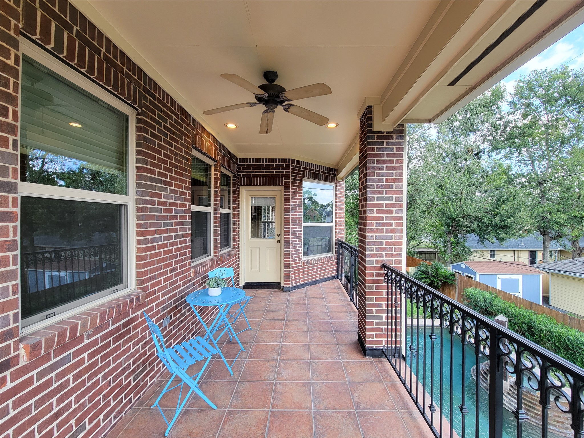 1118 Colonial Street Bellaire, TX 77401 - Photo 31 of 50 This photo showcases a spacious, charming, covered balcony with brick walls and tiled flooring, featuring a ceiling fan and recessed lighting. View of the backyard pool and lush landscaping, providing a perfect space for relaxation.