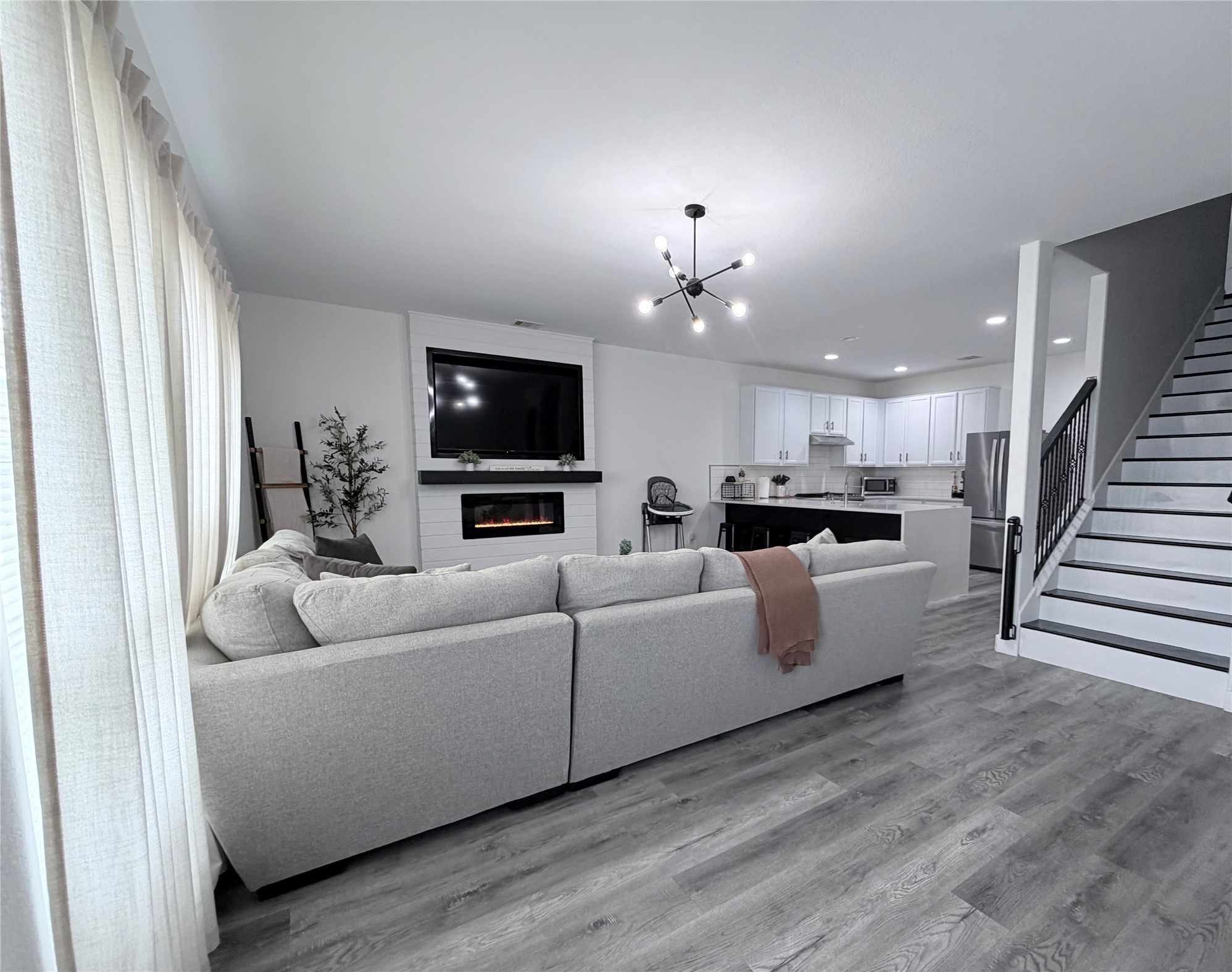 This spacious and modern open-concept living area features a cozy sectional sofa, a sleek fireplace with a mounted TV, and a stylish kitchen with white cabinetry. The light flooring and contemporary lighting add to the airy feel, complemented by a staircase leading to the upper level. Perfect for comfortable living and entertaining.