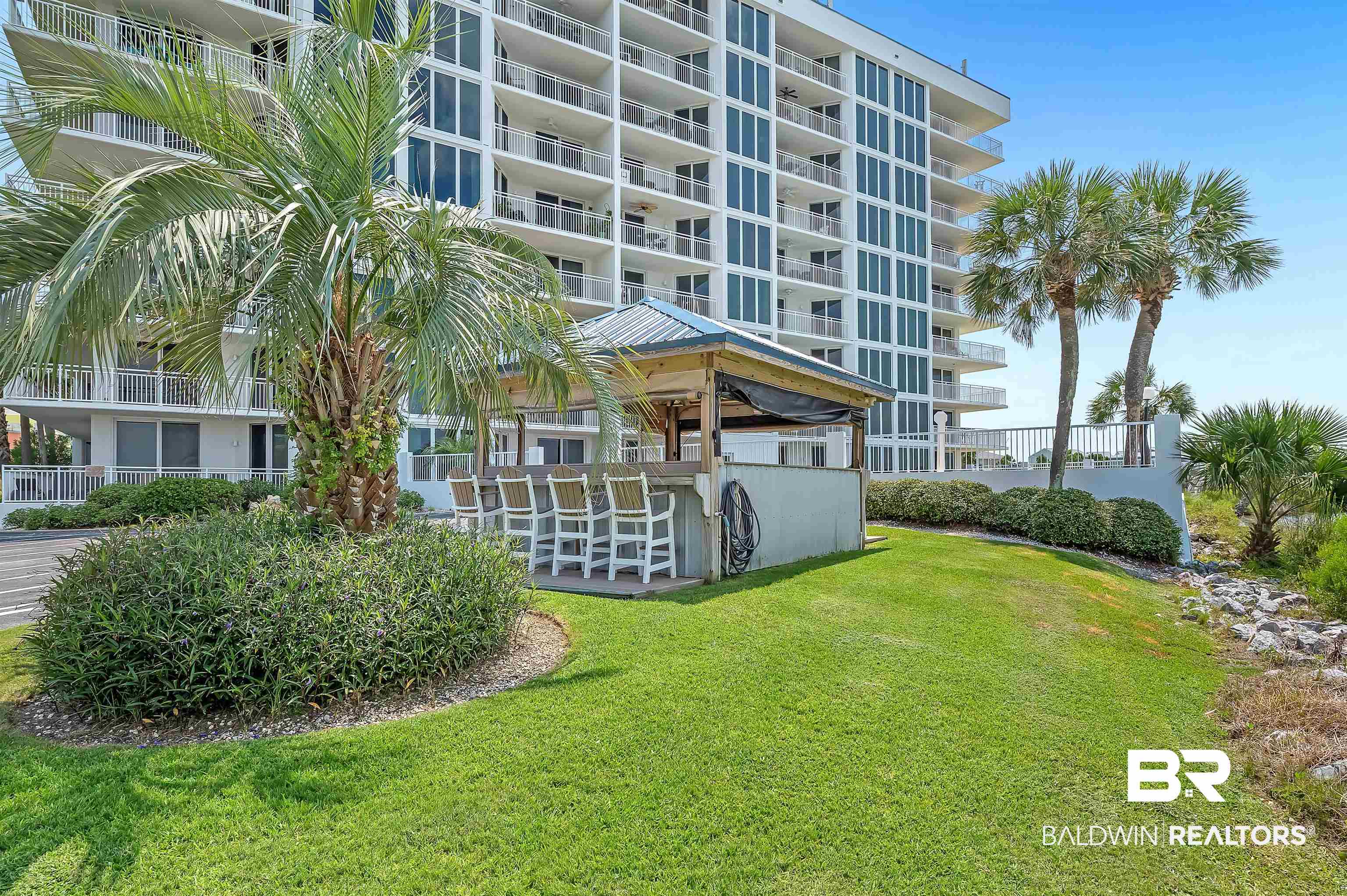 16287 Perdido Key Drive, Unit 105 Pensacola, FL 32507 - Photo 55 of 66 Outdoor bar area with a metal roof and bar seating, surrounded by a manicured lawn and mature palm trees