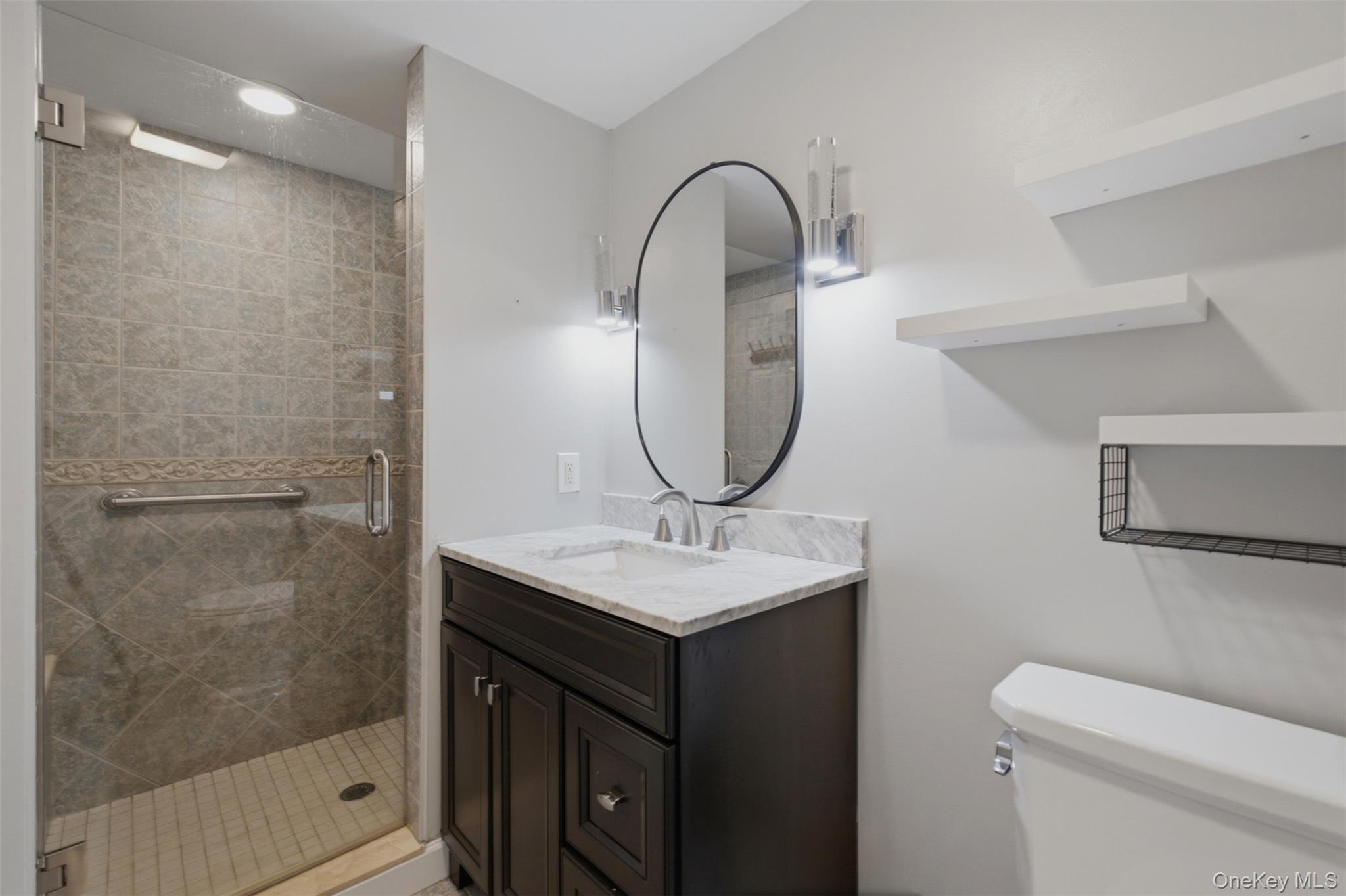 12 Leeside Road Carmel, NY 10512 - Photo 14 of 44 Bathroom featuring a glass-enclosed shower with tile surround, dark wood-finish vanity with a light-toned countertop, oval mirror, and wall-mounted shelving