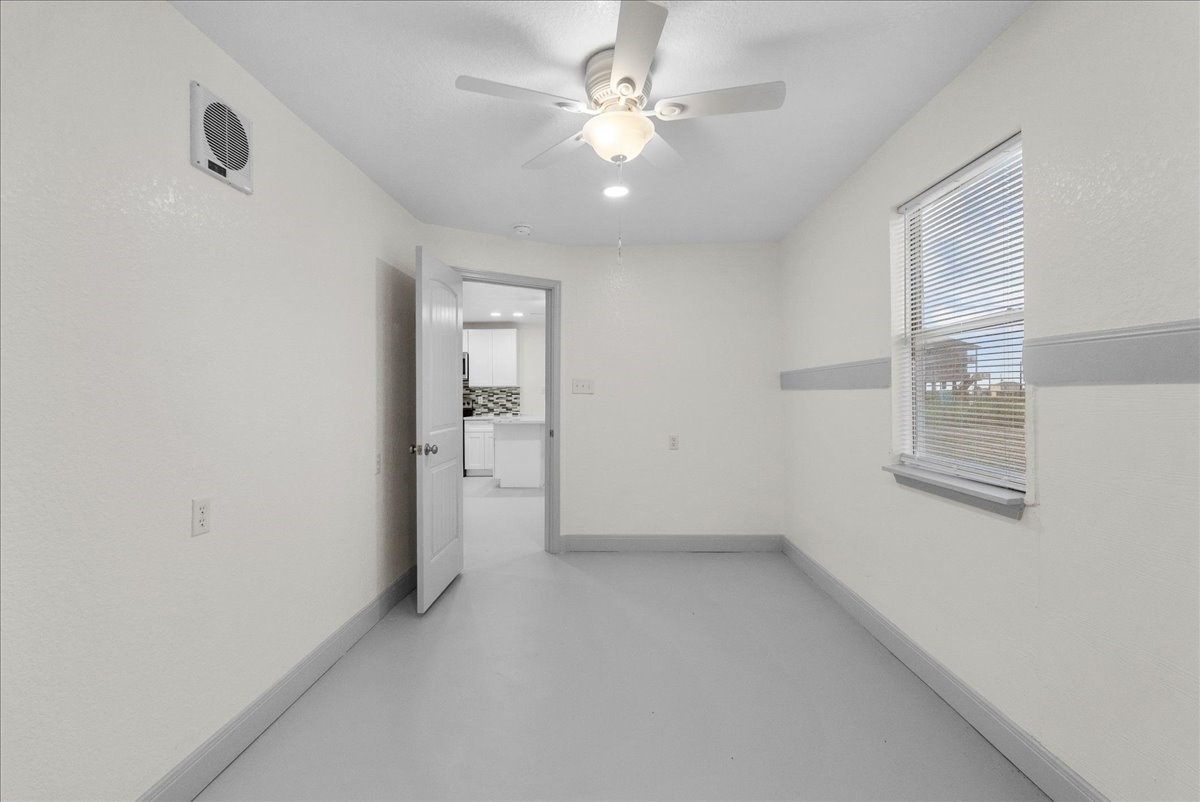 515 Thunder Road Surfside Beach, TX 77541 - Photo 16 of 31 A bright, compact room with neutral walls and gray baseboards, featuring a ceiling fan and a window with blinds. The open door leads to a modern kitchen area with white cabinetry and a mosaic backsplash.