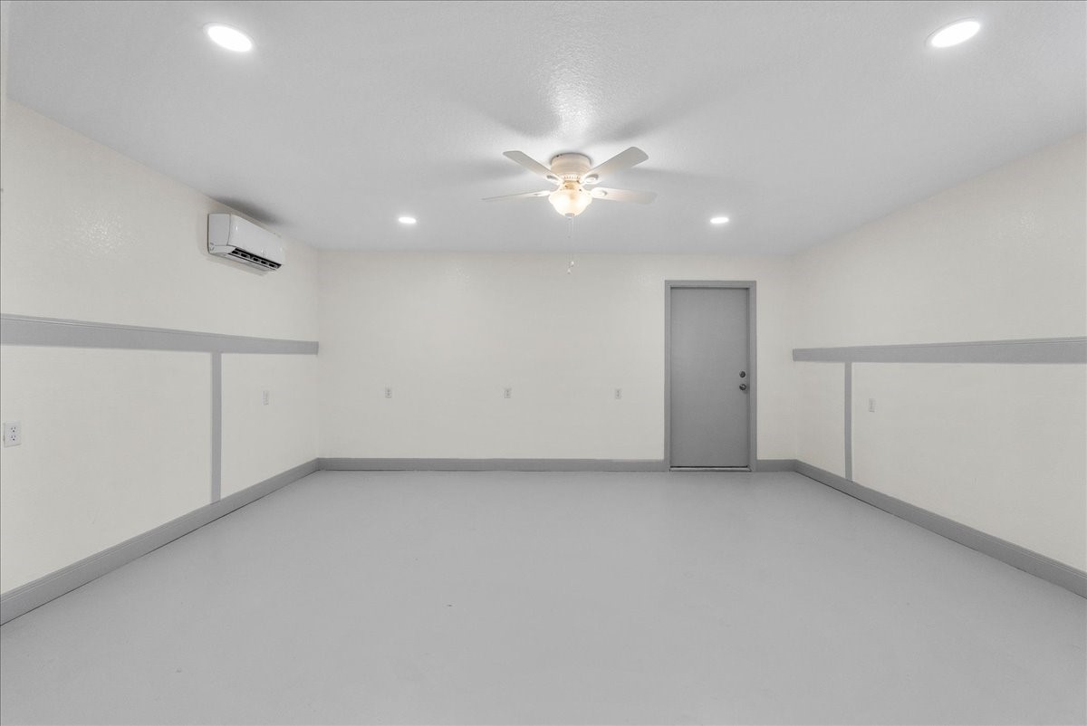 515 Thunder Road Surfside Beach, TX 77541 - Photo 21 of 31 This photo shows a spacious, well-lit room with clean white walls and a gray floor. It features a ceiling fan, recessed lighting, an air conditioning unit, and a single door to a versatile space like a garage or workshop.