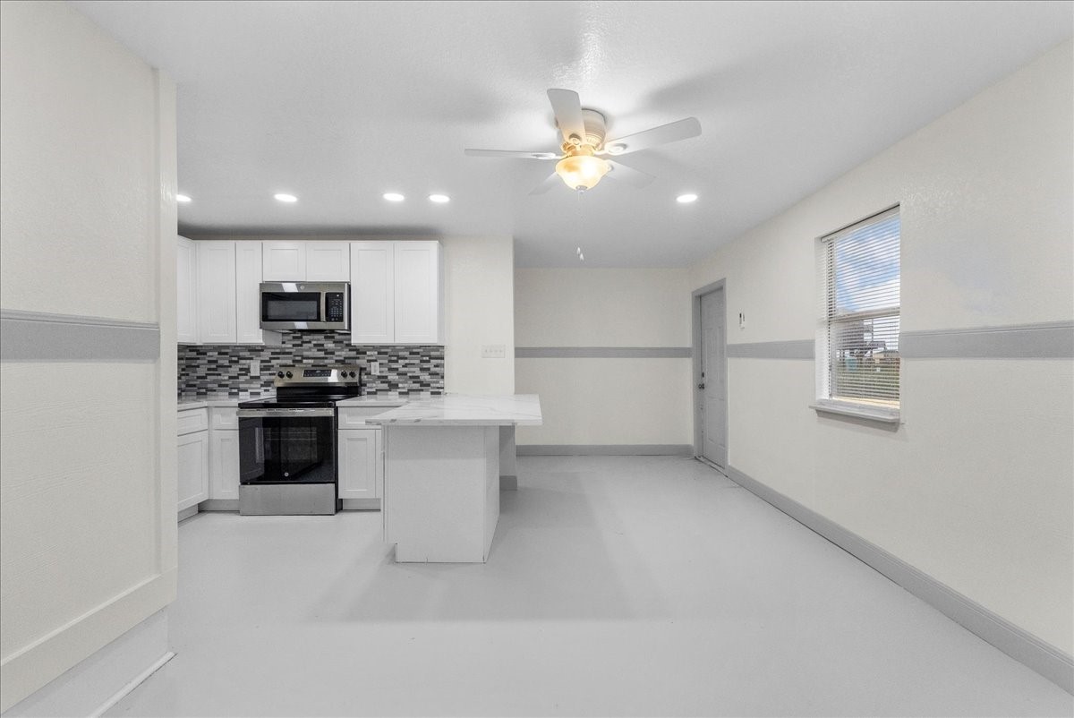 515 Thunder Road Surfside Beach, TX 77541 - Photo 4 of 31 Bright and modern kitchen with white cabinetry, a stylish tile backsplash, and stainless steel appliances. The open layout includes an island and a ceiling fan, with ample natural light from a large window.