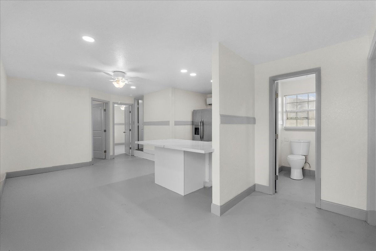 515 Thunder Road Surfside Beach, TX 77541 - Photo 7 of 31 This photo shows a bright, spacious room with light walls and gray trim. It features a ceiling fan, recessed lighting, and a small kitchen area with a counter. There's a bathroom with a shower. Overall, it offers a clean and modern look.