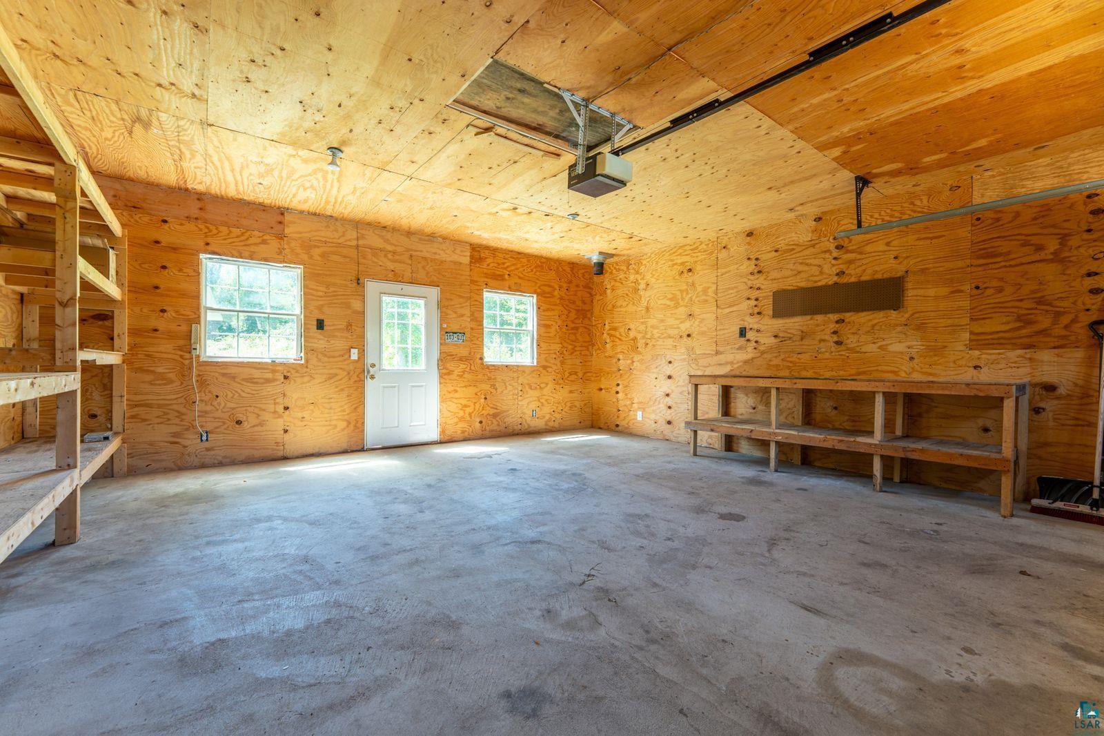 9734 Alango Road Angora, MN 55703 - Photo 19 of 41 Spacious garage featuring plywood-paneled walls and ceiling, a concrete floor, built-in shelving, a workbench, and two windows providing natural light