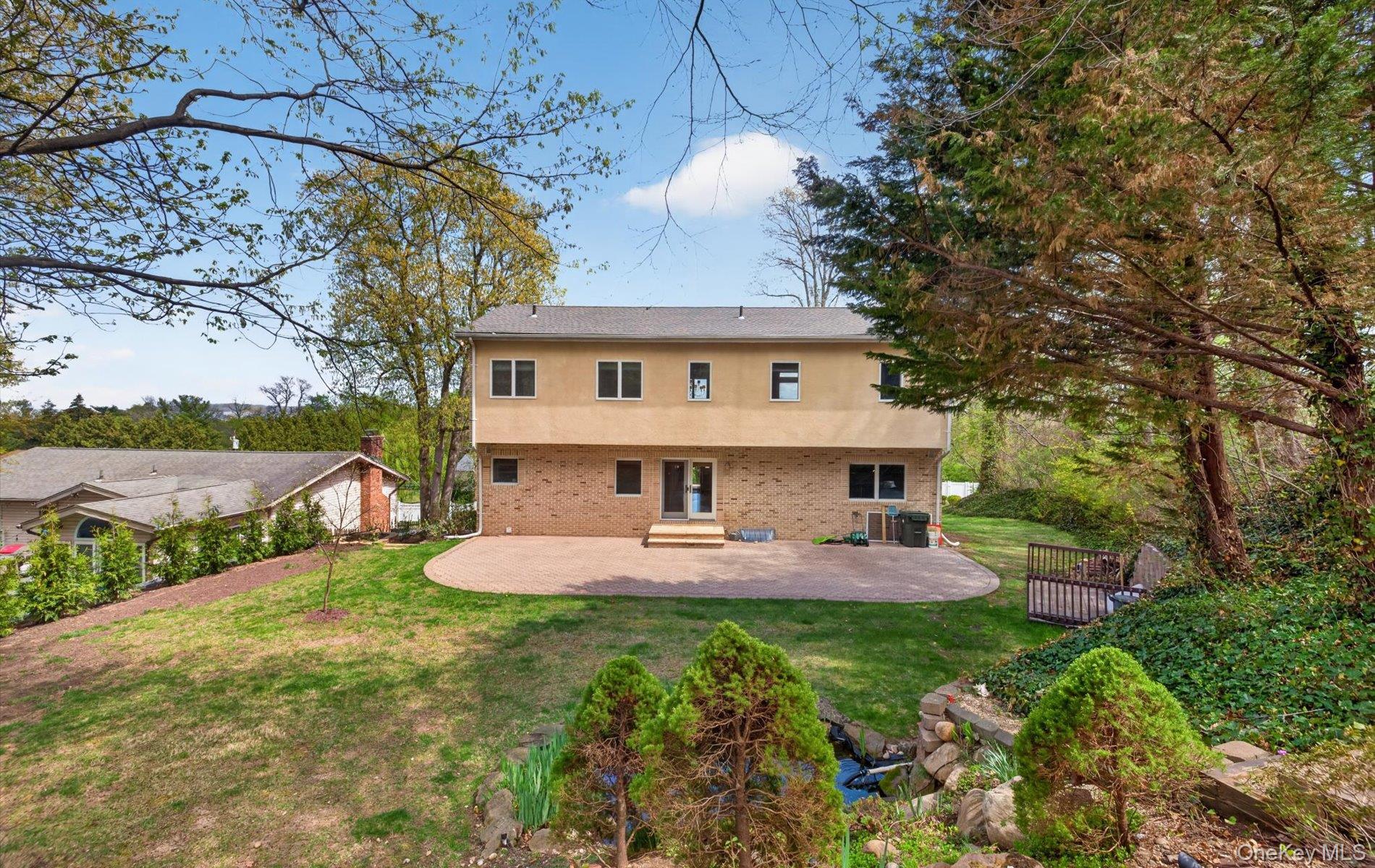 16 East Slope Road Bayville, NY 11709 - Photo 40 of 48 Rear exterior featuring a spacious paver patio, stucco and brick facade, multiple windows, and a landscaped backyard with mature trees