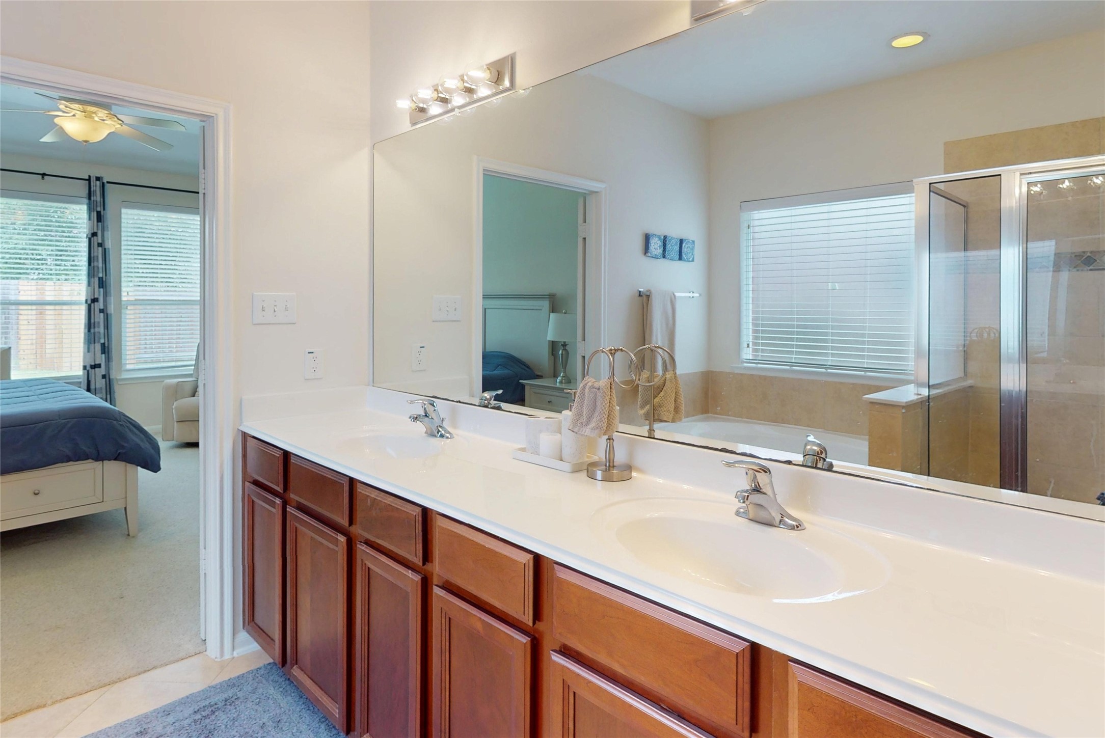 17402 Rainer Valley Lane Humble, TX 77346 - Photo 17 of 33 This bright bathroom features a double vanity with elegant wooden cabinets, a large mirror, and a glass-enclosed shower. It’s connected to a bedroom with ample natural light from large windows.