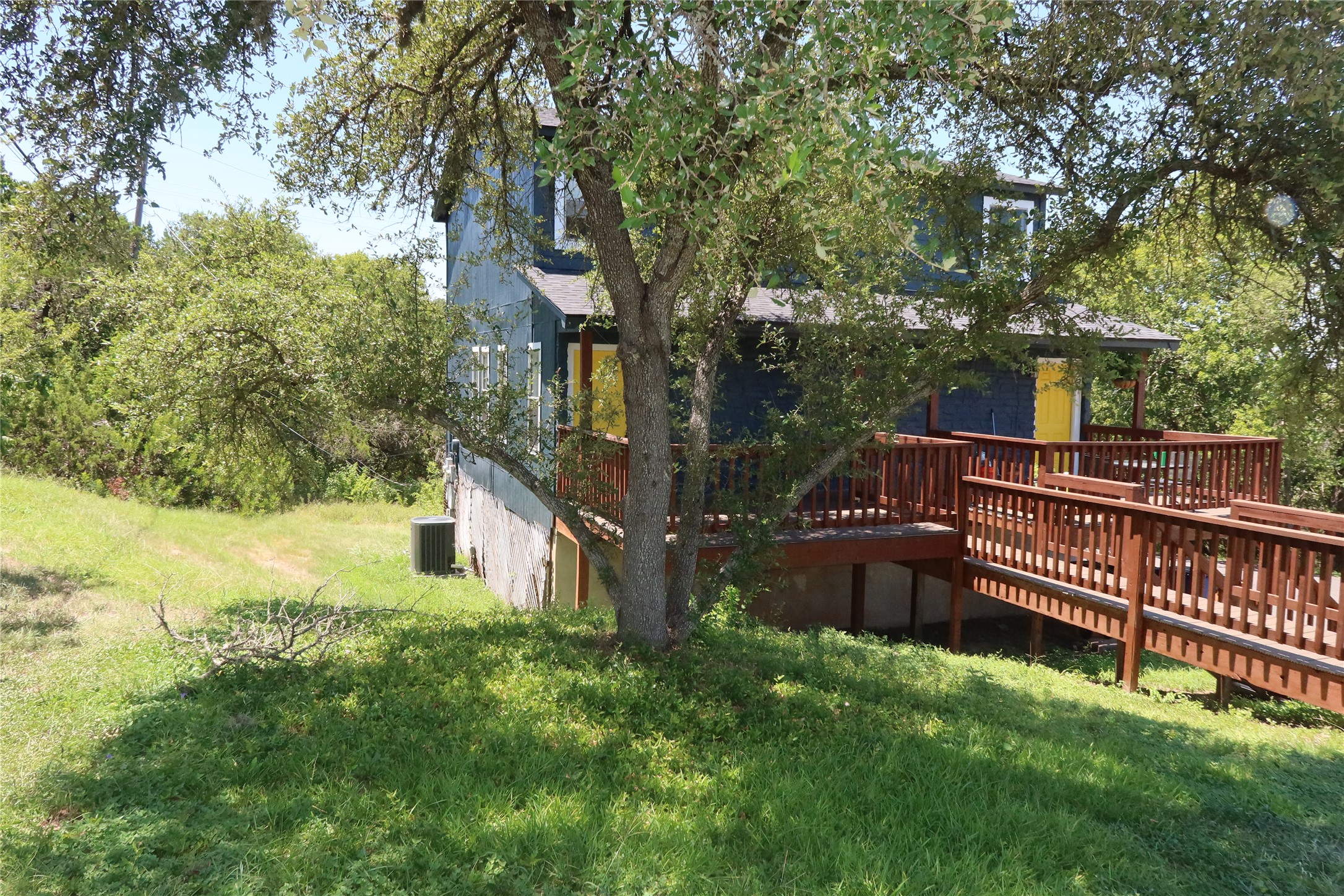 Mature shade trees surround a multi-level wooden deck leading to the entrance.