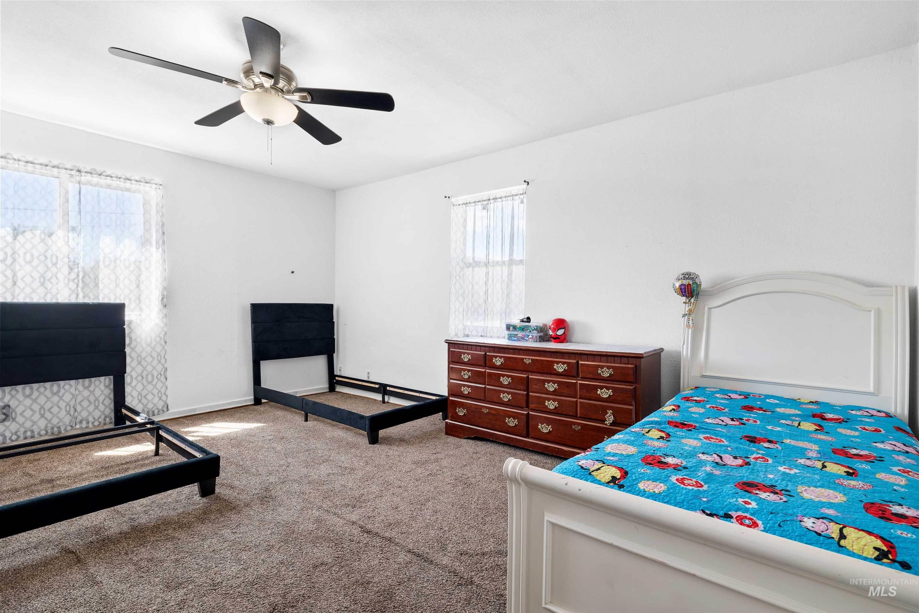 102 East 6th Street Rupert, ID 83350 - Photo 15 of 27 Carpeted room with white walls, two window openings with sheer curtains, a ceiling fan with light fixture, and a wood-finish dresser with multiple drawers