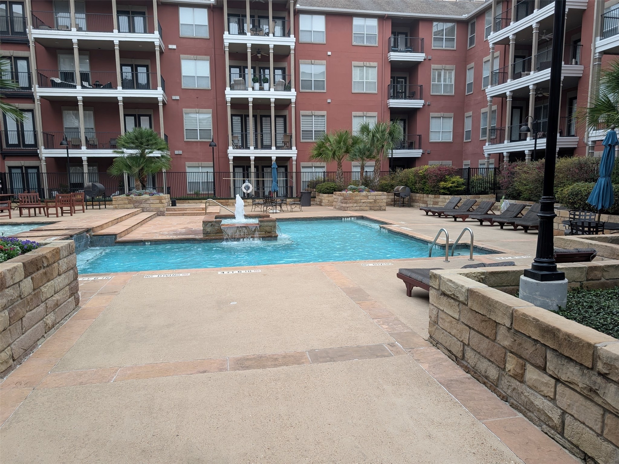 2400 McCue Road, Unit 136 Houston, TX 77056 - Photo 1 of 25 This photo showcases a well-maintained courtyard with a swimming pool and a fountain, surrounded by a multi-story residential building. The area features lounging chairs, palm trees, and a mix of brick and stone accents, creating a relaxing atmosphere.