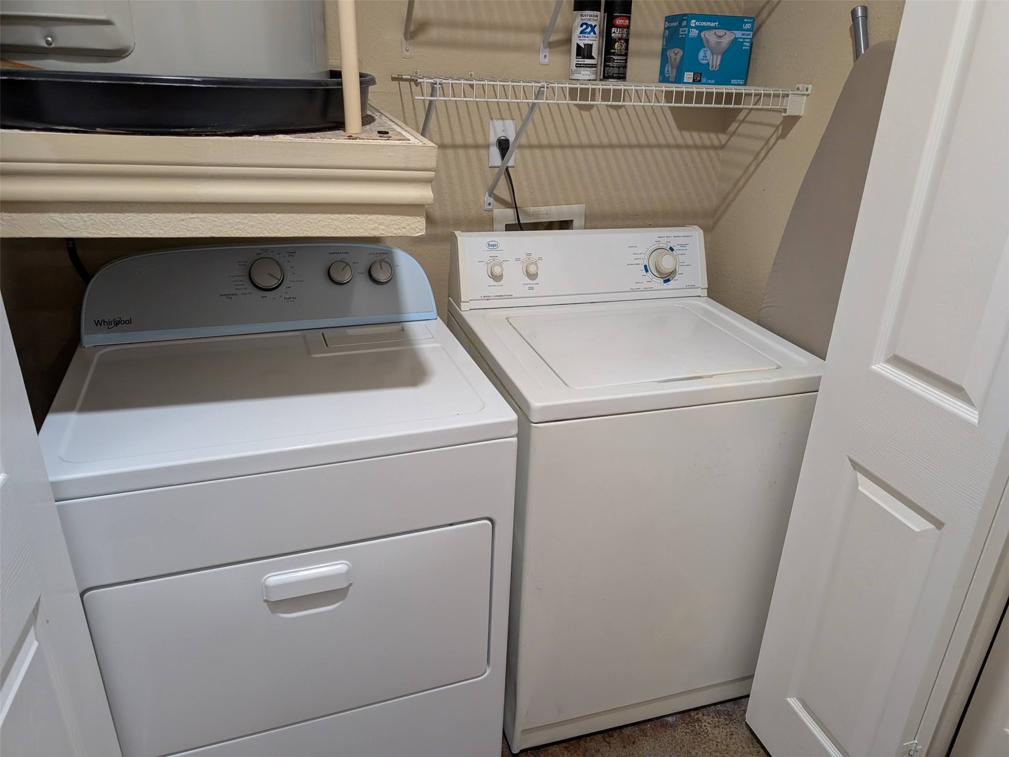 2400 McCue Road, Unit 136 Houston, TX 77056 - Photo 11 of 25 This photo shows a compact laundry area featuring a white washer and dryer set. The space includes a wire shelf above for storage and an ironing board tucked neatly to the side, offering practical utility in a small area.
