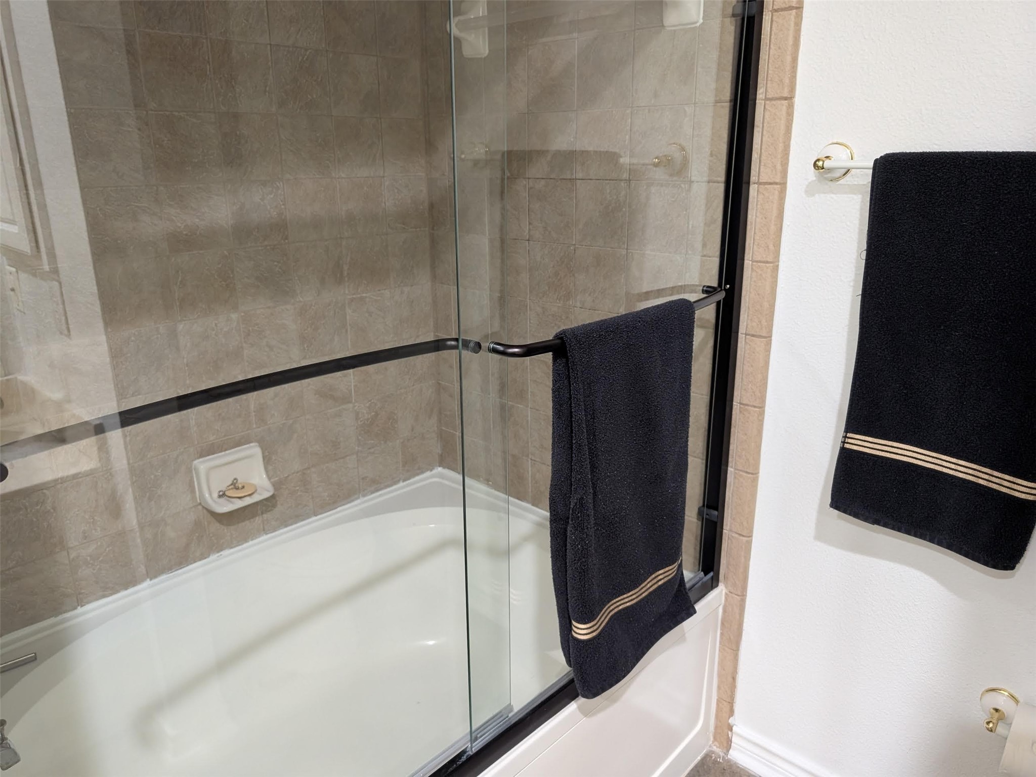 2400 McCue Road, Unit 136 Houston, TX 77056 - Photo 14 of 25 This bathroom features a clean, modern design with a glass-enclosed bathtub and shower, beige tiled walls, and sleek black towel accents.