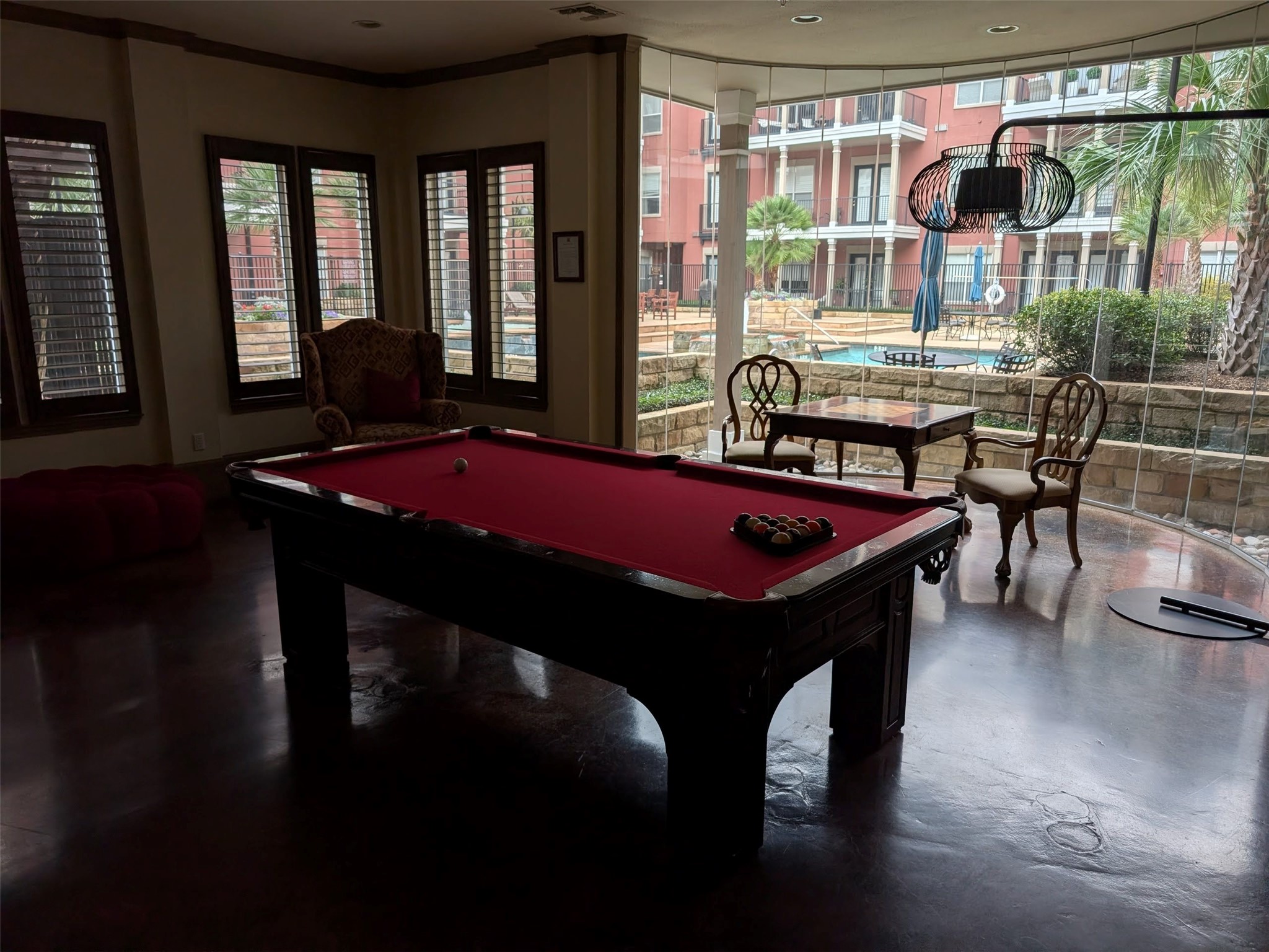 2400 McCue Road, Unit 136 Houston, TX 77056 - Photo 20 of 25 This photo showcases a stylish recreation room with a red-felt pool table, elegant seating, and large windows offering a view of a pool and patio area outside. The space is well-lit and inviting, ideal for entertainment and relaxation.