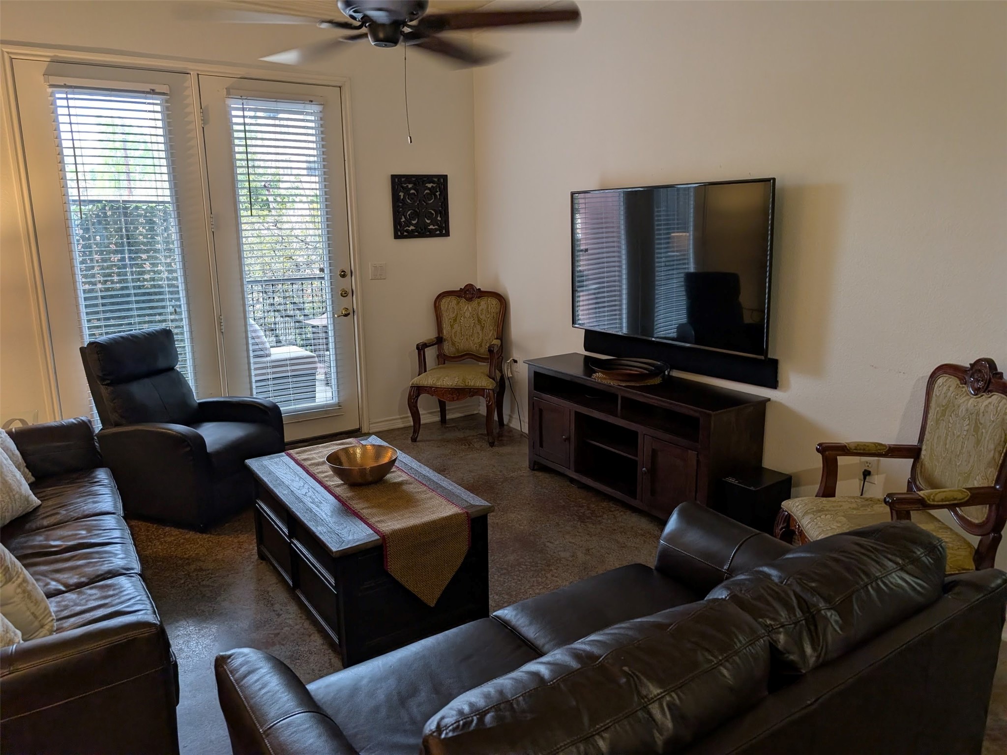 2400 McCue Road, Unit 136 Houston, TX 77056 - Photo 2 of 25 Cozy living room with a comfortable seating arrangement, featuring a large TV on a wooden stand. Natural light filters through glass doors with blinds, leading to an outdoor area, and a ceiling fan adds to the comfort.
