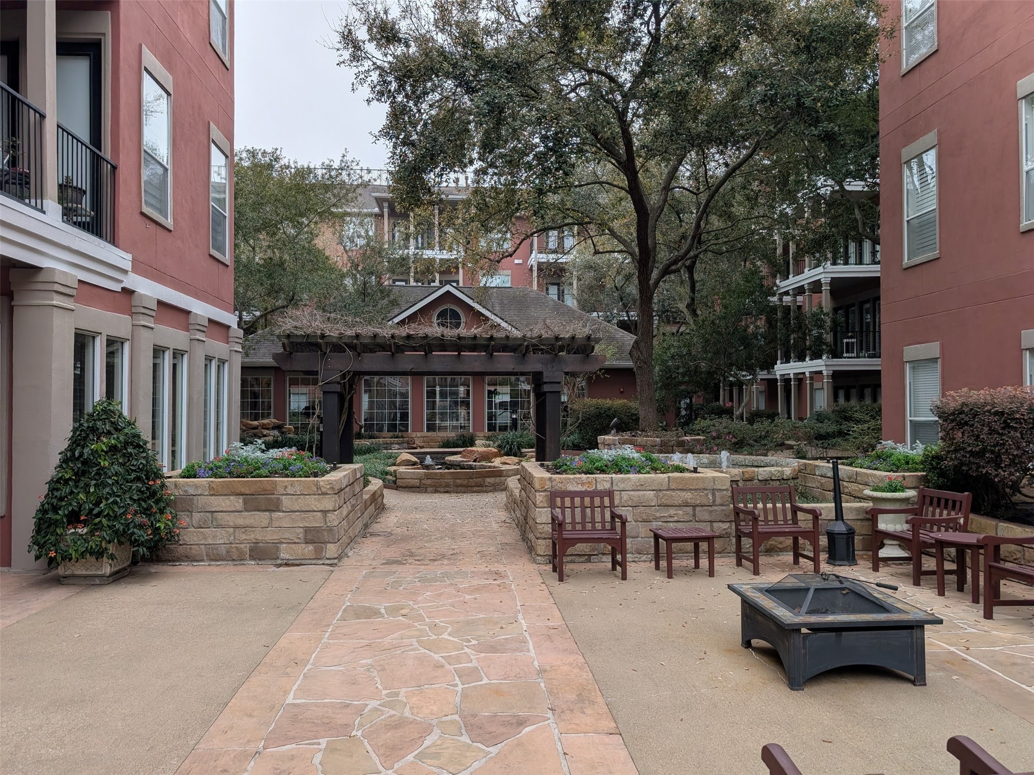 2400 McCue Road, Unit 136 Houston, TX 77056 - Photo 23 of 25 This photo showcases a charming courtyard within a residential complex, featuring a paved pathway, lush landscaping, and comfortable seating areas. The space is framed by red-brick buildings and includes a pergola, offering a serene outdoor retreat.