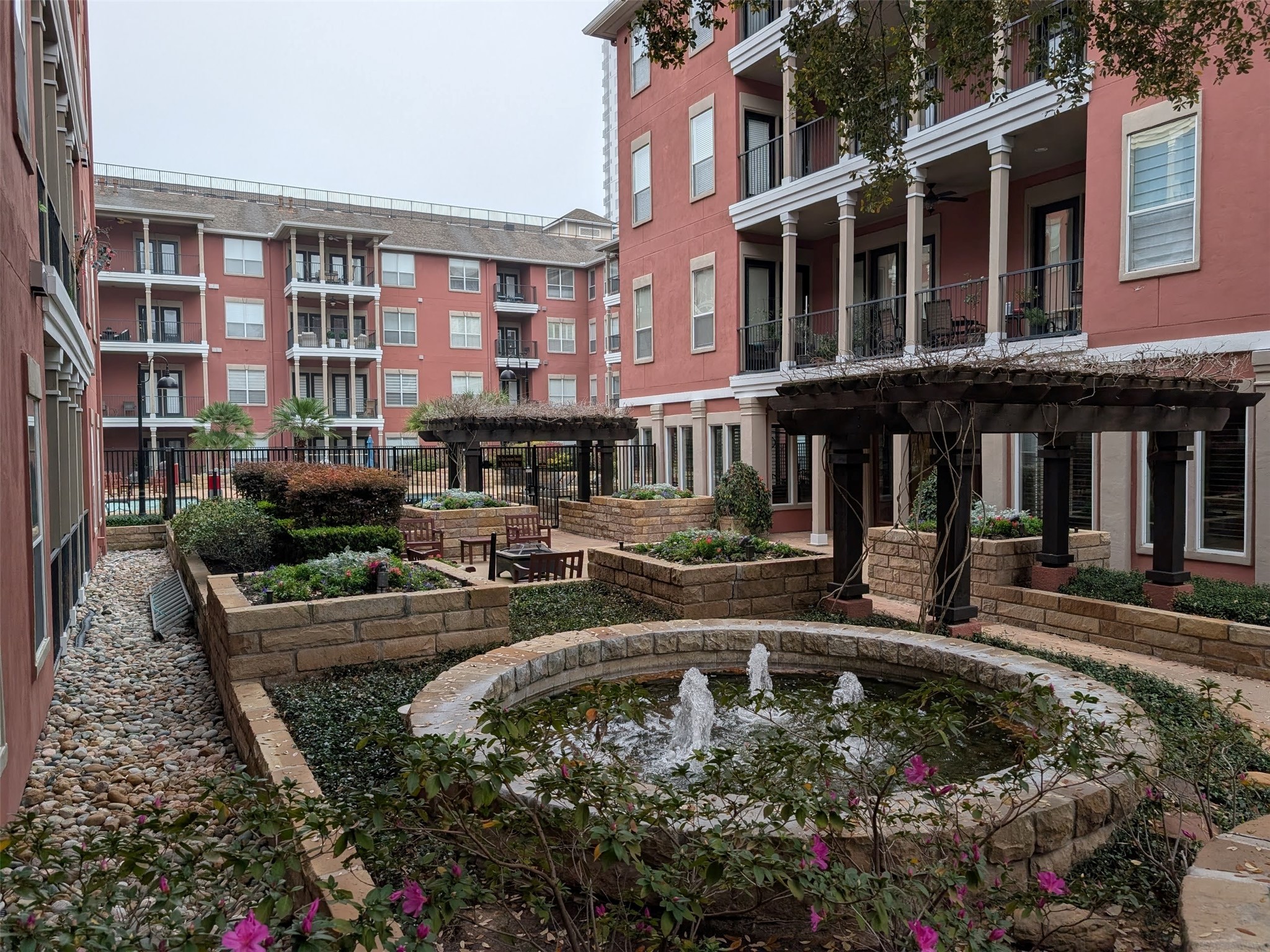 2400 McCue Road, Unit 136 Houston, TX 77056 - Photo 24 of 25 This photo showcases a charming apartment courtyard with landscaped gardens, a central circular fountain, and outdoor seating areas. The surrounding buildings feature balconies, adding to the inviting, community-focused atmosphere.