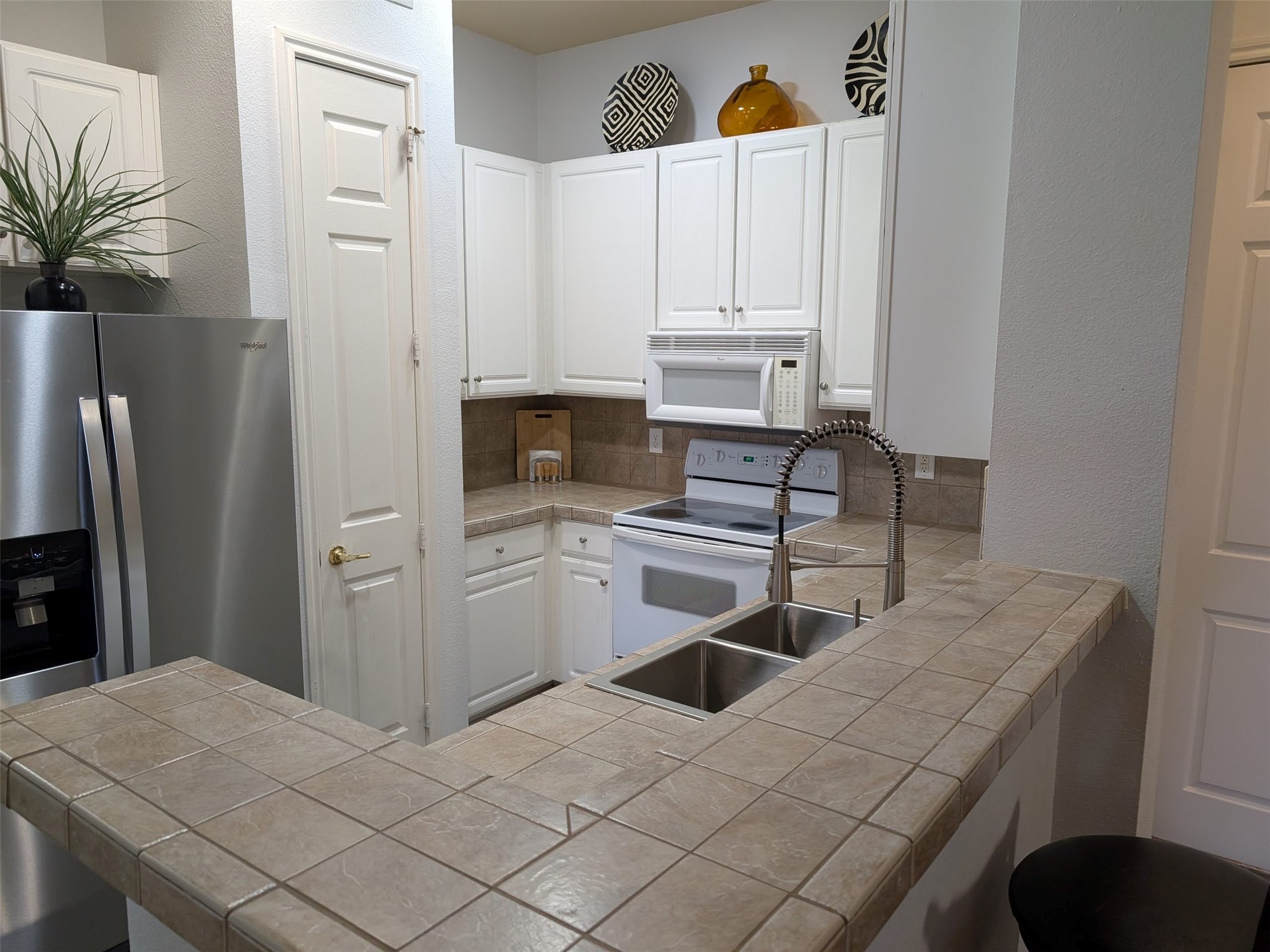 2400 McCue Road, Unit 136 Houston, TX 77056 - Photo 5 of 25 This kitchen features white cabinetry, a tile countertop, and backsplash, with stainless steel appliances, including a fridge. It has a built-in oven and microwave, a double sink with a modern faucet, and decorative accents above the cabinets. Perfect for a cozy cooking space.
