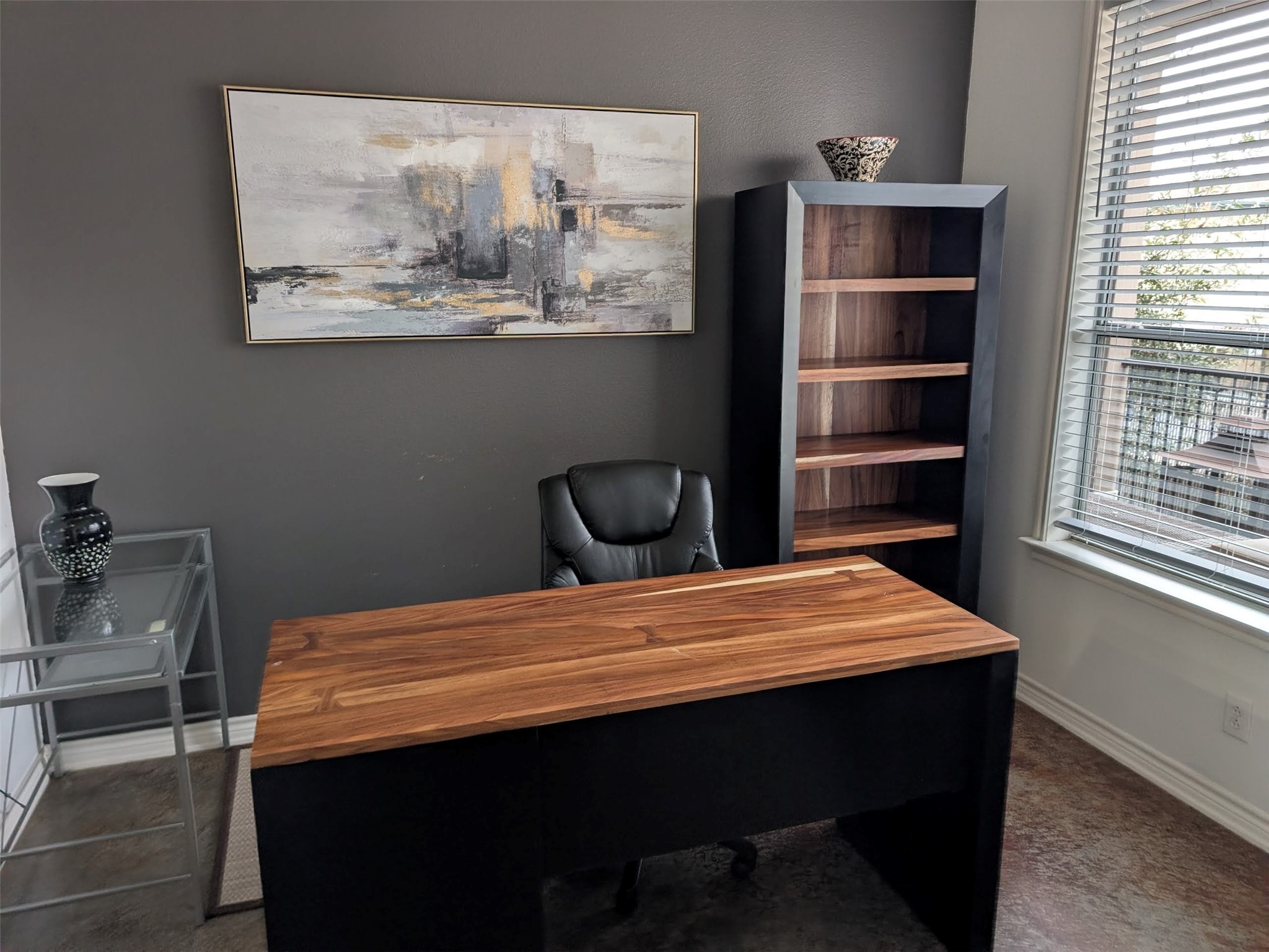 2400 McCue Road, Unit 136 Houston, TX 77056 - Photo 7 of 25 This photo showcases a modern home office with a sleek wooden desk and a black leather chair. There's a large abstract painting on a gray accent wall, a tall bookshelf, and natural light streaming in through a window with blinds. A glass side table with decorative vases adds a stylish touch.