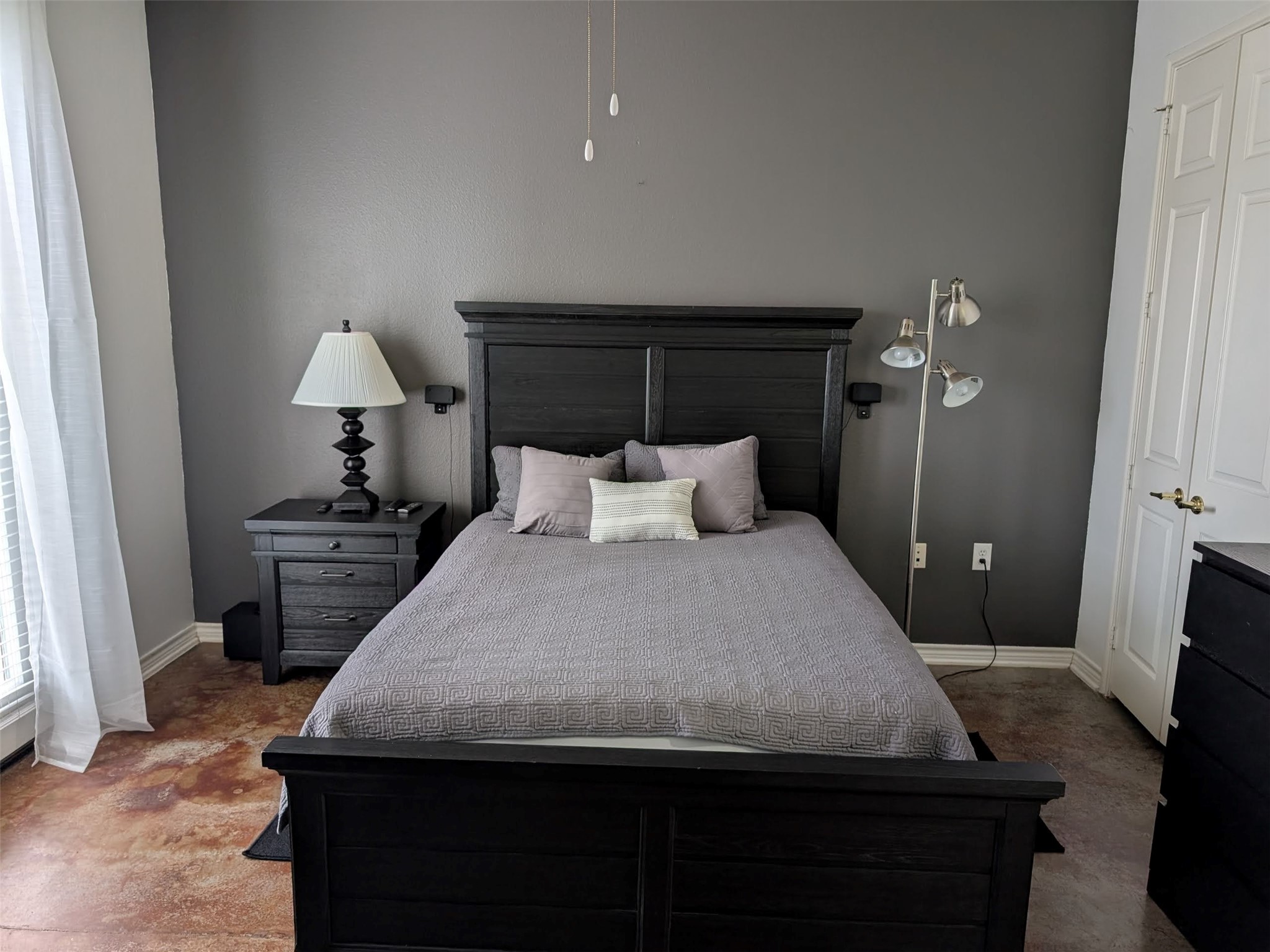 2400 McCue Road, Unit 136 Houston, TX 77056 - Photo 8 of 25 This bedroom features a modern, minimalist design with a dark wooden bed frame and matching nightstand. It has a neutral color palette with gray walls and a light gray bedspread. Natural light filters through the window with sheer curtains, giving the room a bright and airy feel.