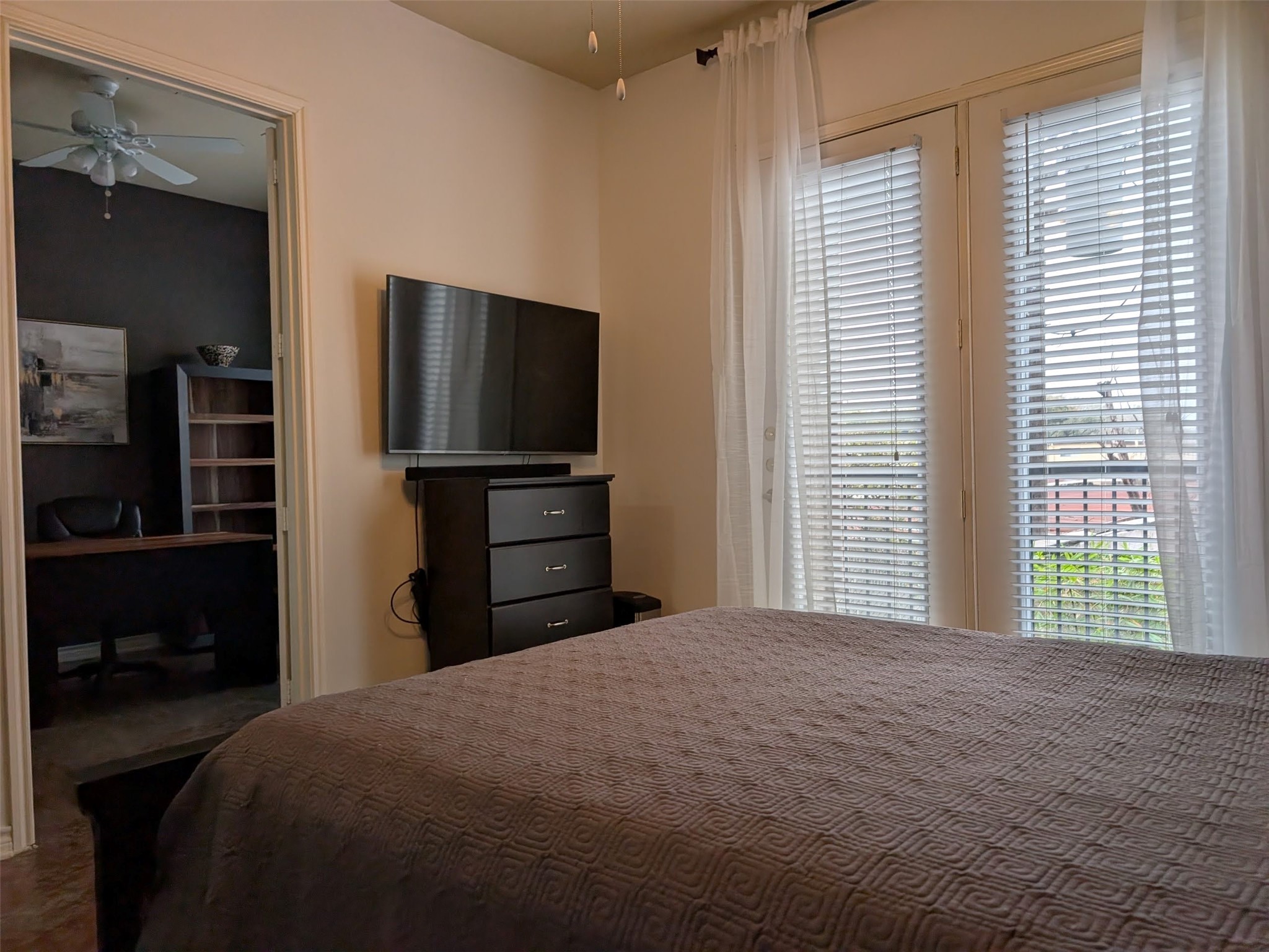 2400 McCue Road, Unit 136 Houston, TX 77056 - Photo 10 of 25 The bedroom features a cozy setup with a mounted TV, dresser, and large windows with blinds and sheer curtains allowing natural light. An adjacent office space is visible through an open doorway, offering a functional work area with a ceiling fan and shelving.
