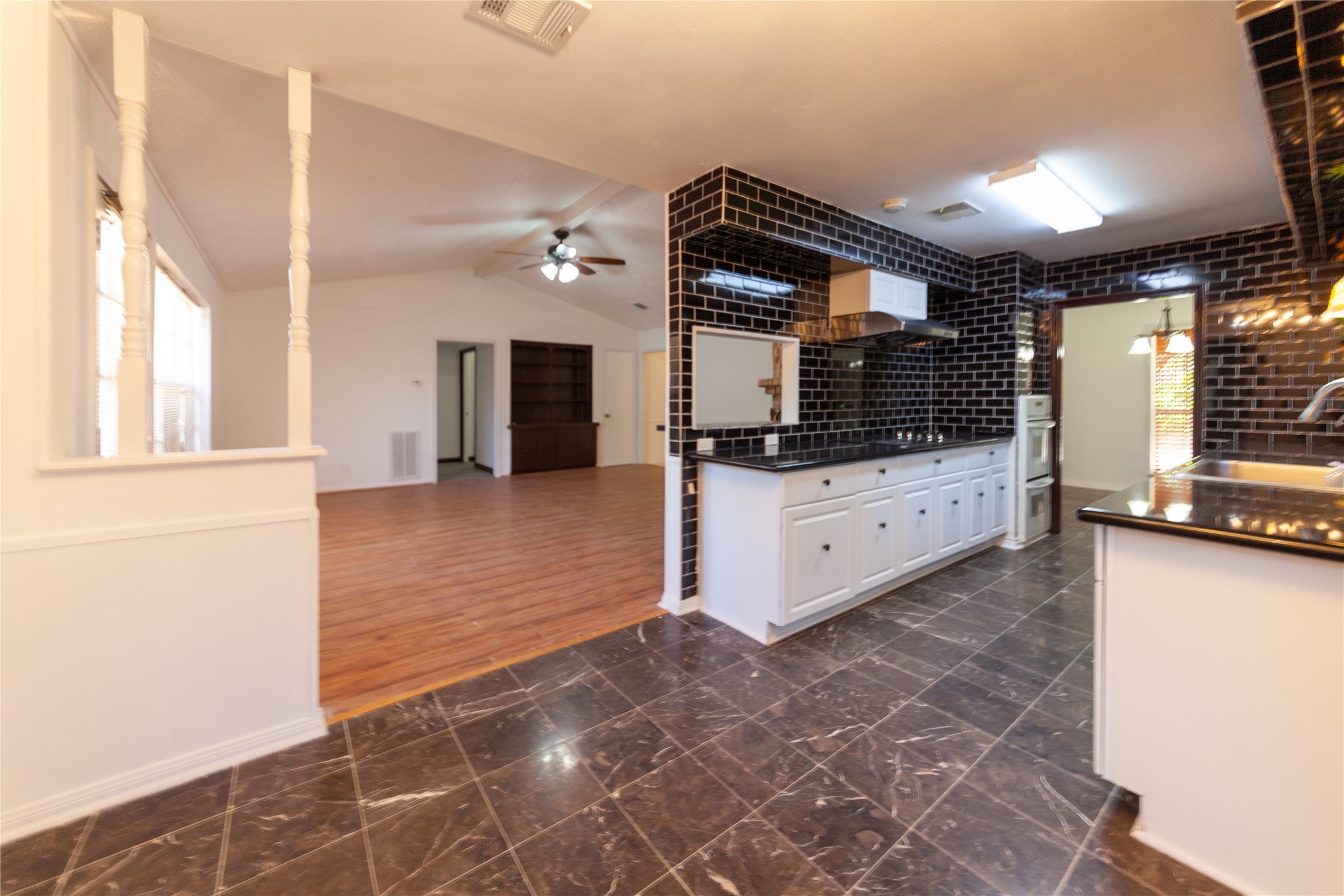 8826 Roos Road Houston, TX 77036 - Photo 9 of 42 An open-concept space featuring a kitchen with sleek black tile walls and countertops, white cabinetry, and a dark tiled floor. The adjacent area has warm wood flooring, a ceiling fan, and built-in shelving, creating a seamless flow between the kitchen and living space.