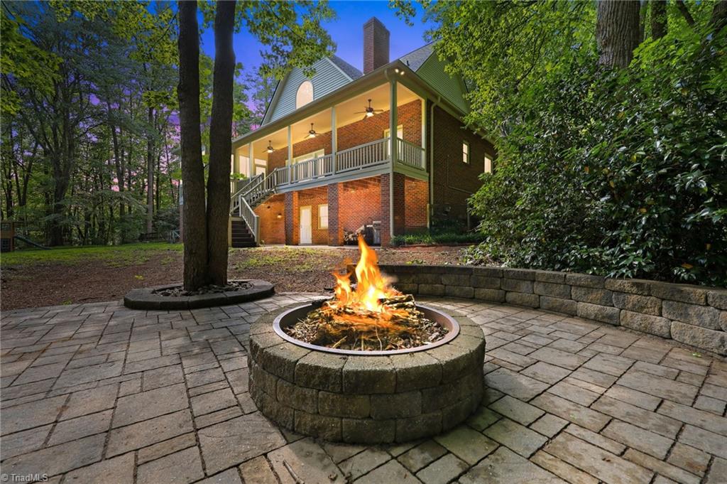 8861 Boylston Road Colfax, NC 27235 - Photo 39 of 50 Gather with family and friends around the custom paver patio and built-in fire pit, creating the perfect setting for cool Carolina evenings. Framed by mature hardwoods and overlooking the beautifully illuminated rear elevation, this private outdoor retreat feels like your own backyard getaway. With multiple outdoor living spaces—including the expansive covered porch, screened porch, and spacious patio—this home was designed for year-round entertaining and making lasting memories. (Enhanced Twilight Image)