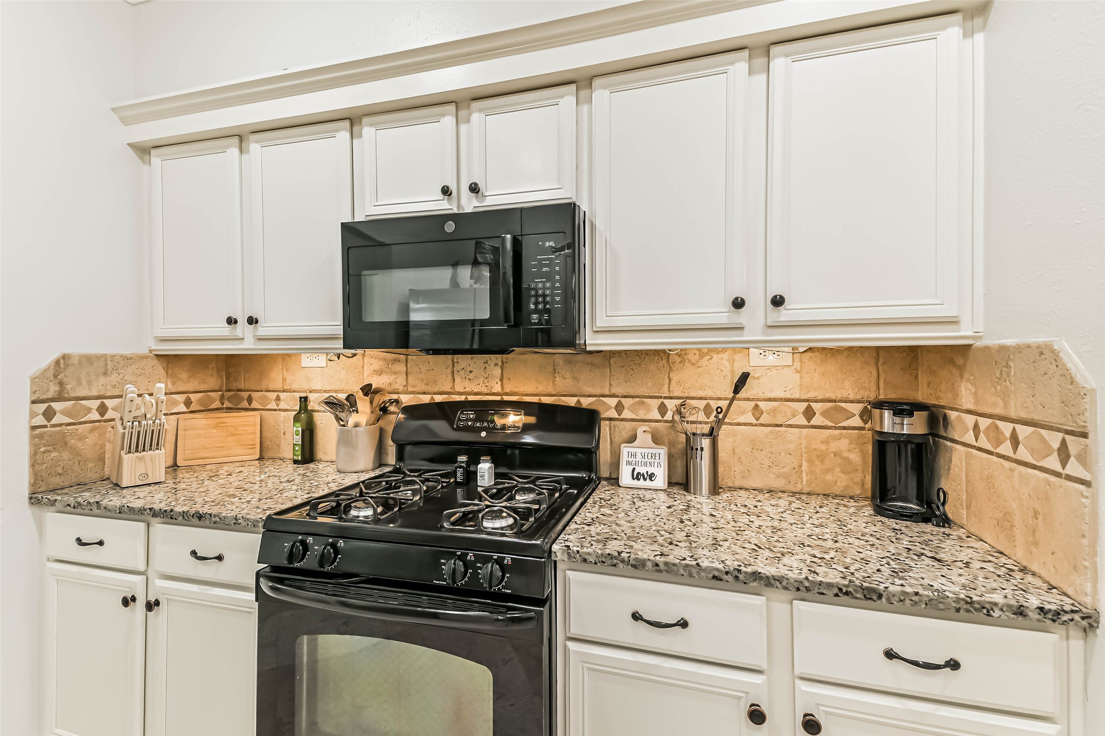 22538 Goss Spring Court Spring, TX 77373 - Photo 10 of 36 This kitchen features white cabinetry with a granite countertop and a stone tile backsplash. It includes a gas stove, built-in microwave, and a sleek black appliance suite, perfect for modern cooking needs.