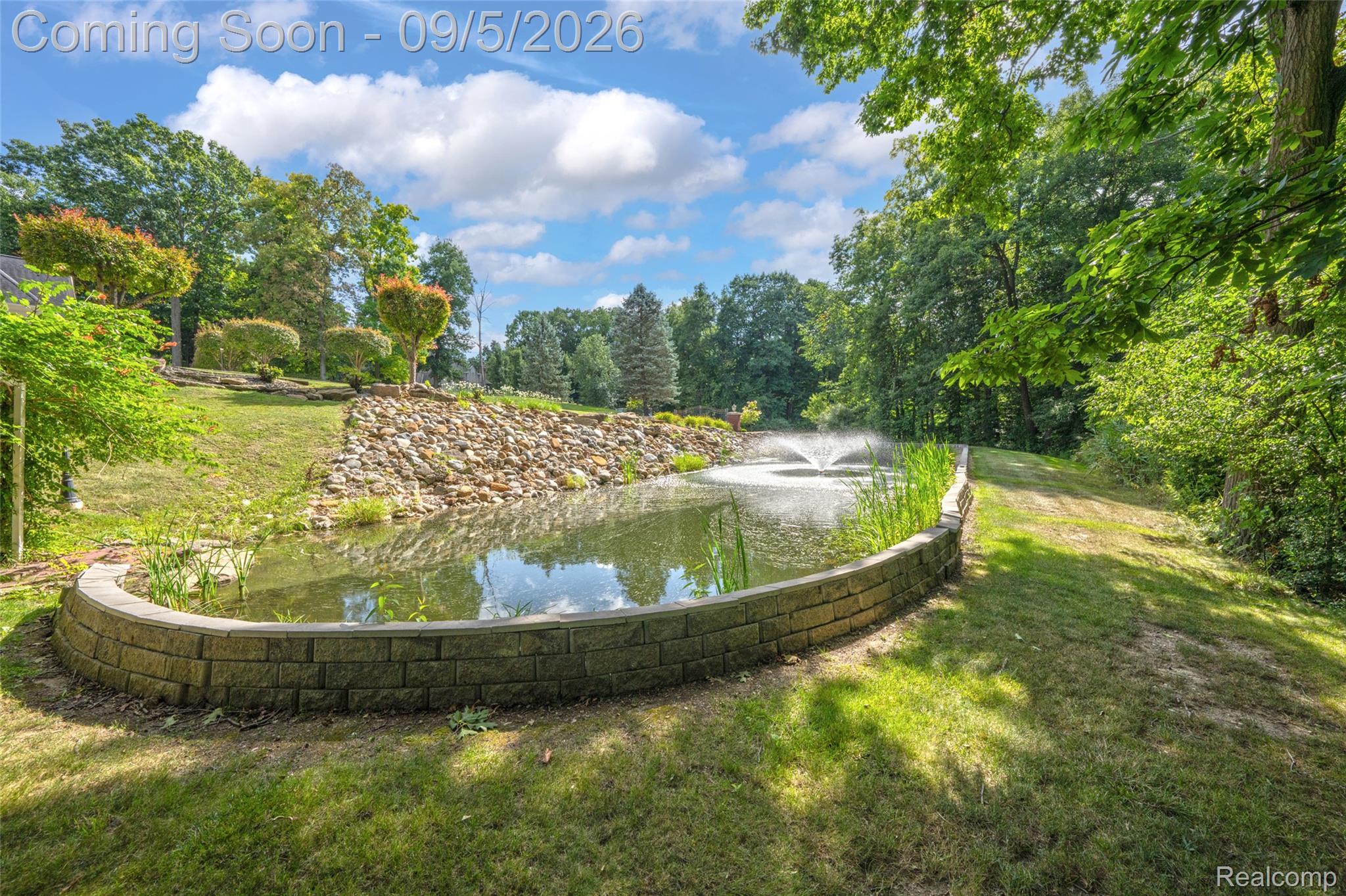2785 Cranbrook Ridge Court Rochester, MI 48306 - Photo 88 of 97 Water feature with a fountain, stone retaining wall, natural rock landscaping, mature trees, and expansive grassy areas