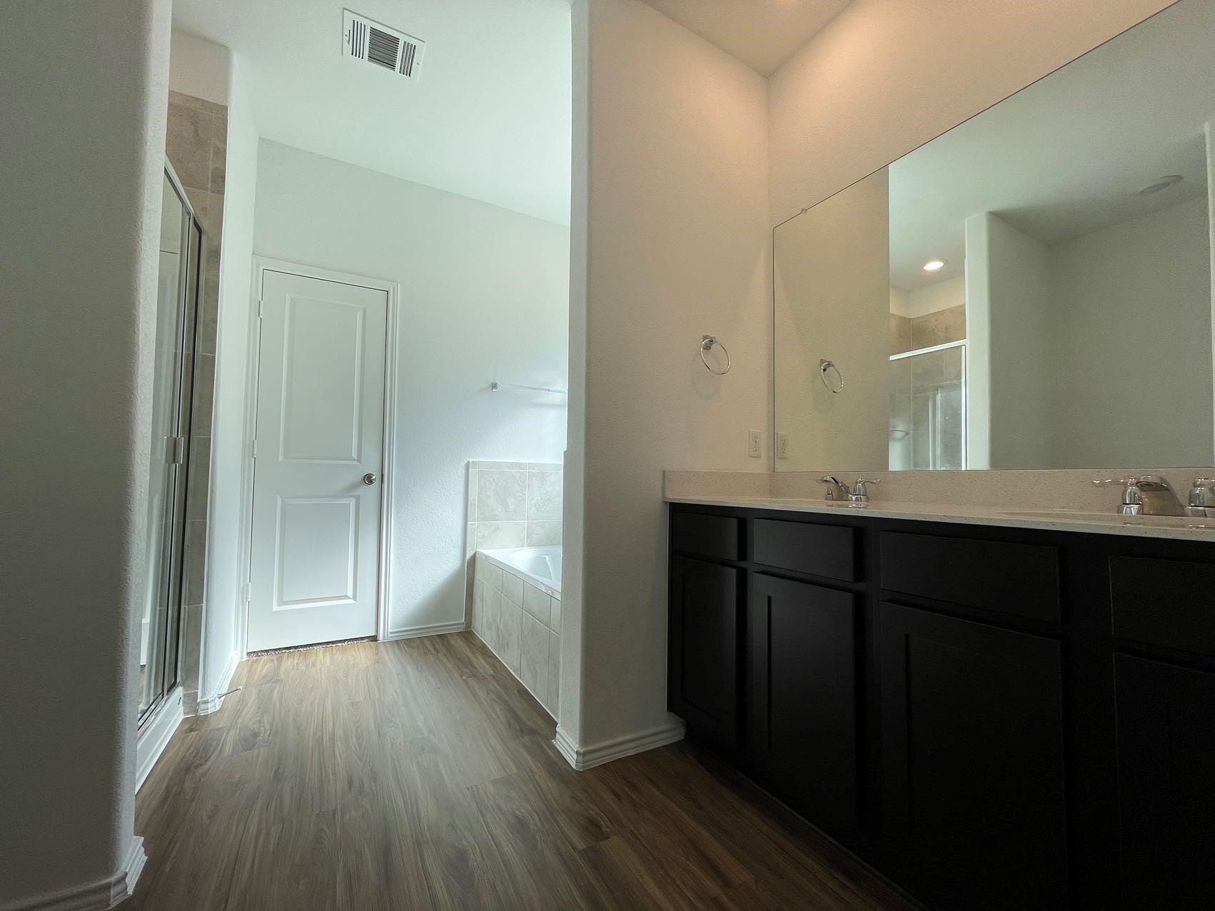 1625 Woolsey Way Leander, TX 78641 - Photo 12 of 17 Bathroom featuring a double vanity with dark cabinetry, a large mirror, a separate shower with a glass enclosure, a bathtub with tiled surround, and wood-look flooring