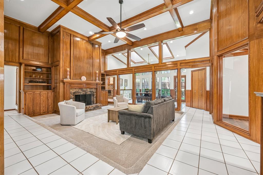 925 Spring Brook Drive Allen, TX 75002 - Photo 5 of 30 Wood-paneled living space featuring a fireplace with a brick surround, built-in shelving, white tile flooring, and a beamed ceiling