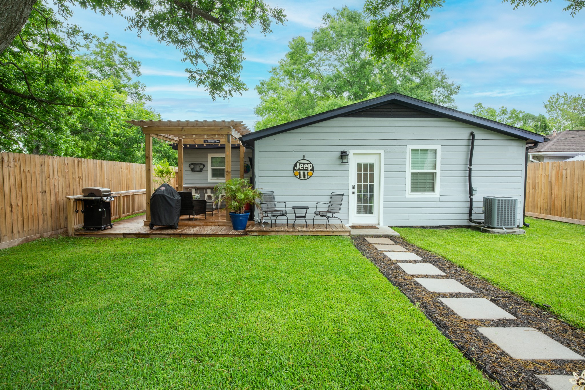 This backyard features a well-maintained lawn, a cozy patio area with outdoor seating, and a grill, perfect for entertaining. The light gray house's rear entrance is accessible via a stone path, and the yard is enclosed by a wooden fence for added privacy.