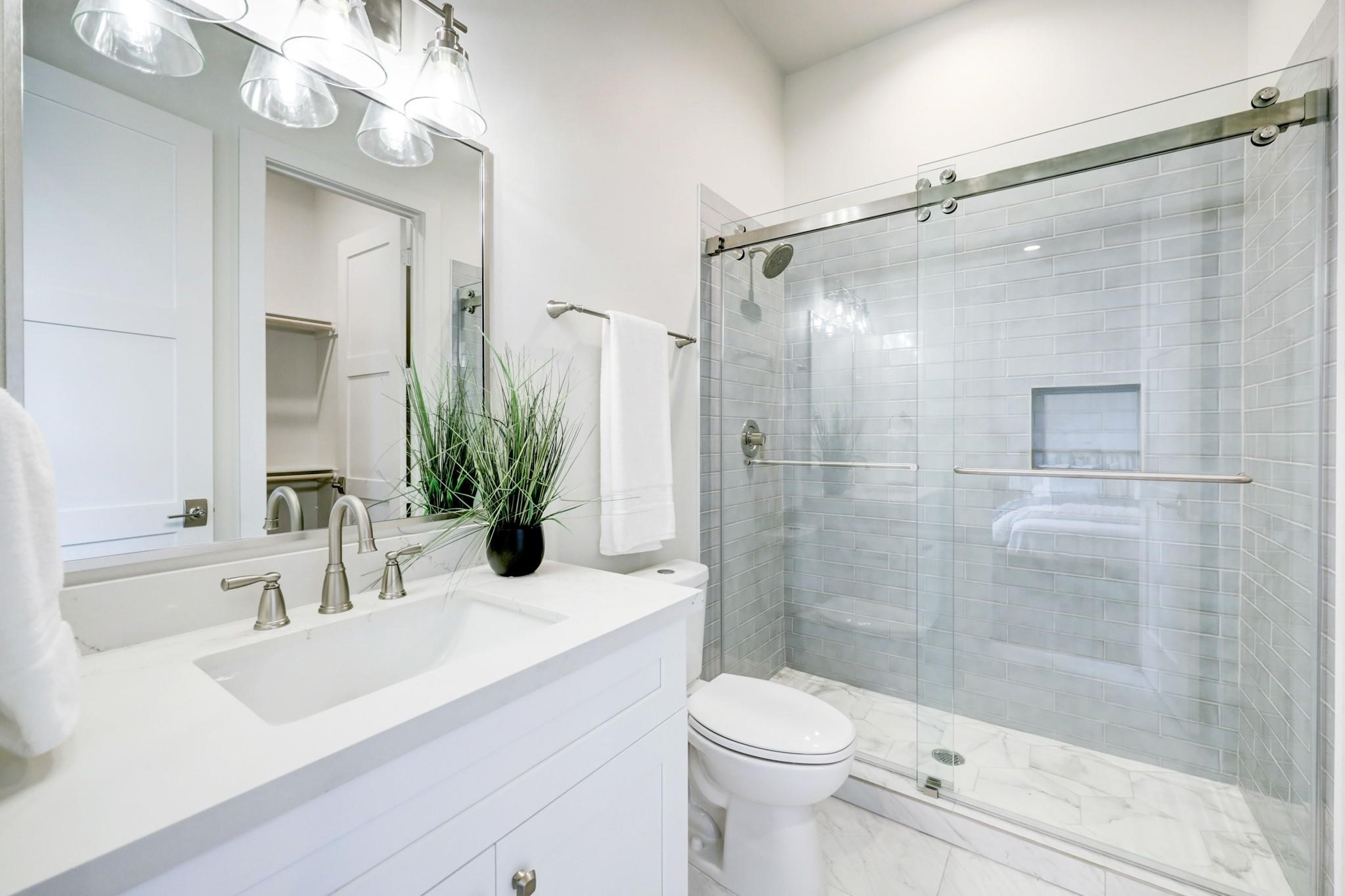 3827 Arlington Street Houston, TX 77018 - Photo 34 of 39 Bathroom features a sleek, white vanity with a stylish faucet and ample counter space. It includes a glass-enclosed shower with elegant subway tiles and a built-in shelf. The space is bright, with contemporary lighting and clean lines, creating a fresh and inviting atmosphere.