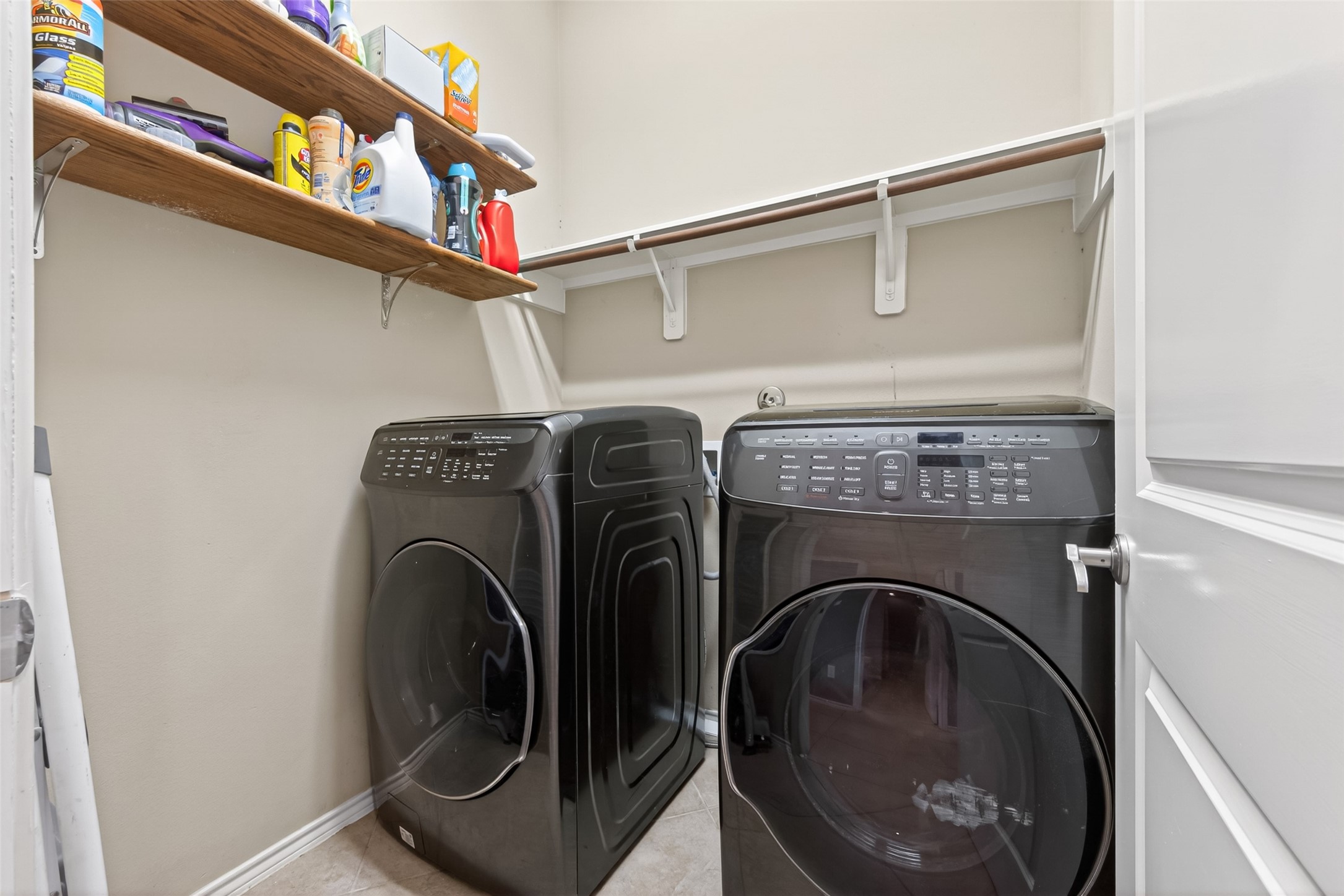 27443 Rebecca Field Lane Spring, TX 77386 - Photo 25 of 31 This photo shows a compact laundry room with a modern washer and dryer. It features overhead shelves for storage, providing space for detergents and other supplies. The room has a clean, functional design with neutral walls and tile flooring.