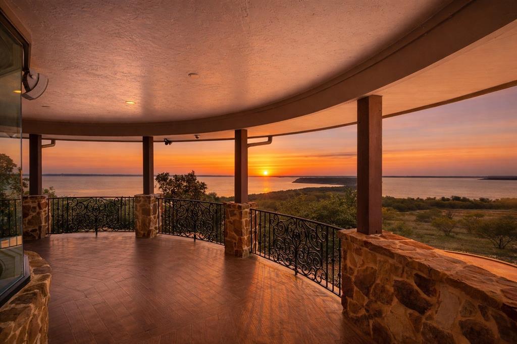 1180 McReynolds Road Sanger, TX 76266 - Photo 31 of 40 Curved covered balcony featuring a wood-finish flooring, stone pillars, and ornate wrought iron railings overlooking a prominent body of water and distant land formations