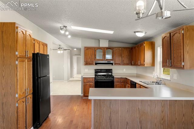 34535 Big Springs Road Yoder, CO 80864 - Photo 8 of 34 Kitchen with black appliances, under cabinet range hood, vaulted ceiling, a peninsula, and brown cabinetry