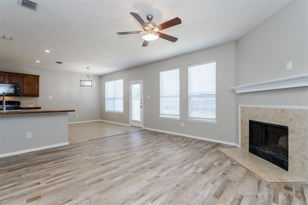 203 Arabian Road Waxahachie, TX 75165 - Photo 8 of 24 Spacious open-concept living area featuring wood-finish flooring, a ceiling fan, multiple windows with blinds, a fireplace with a stone surround and mantel, and an adjacent kitchen with wood cabinetry and a breakfast bar