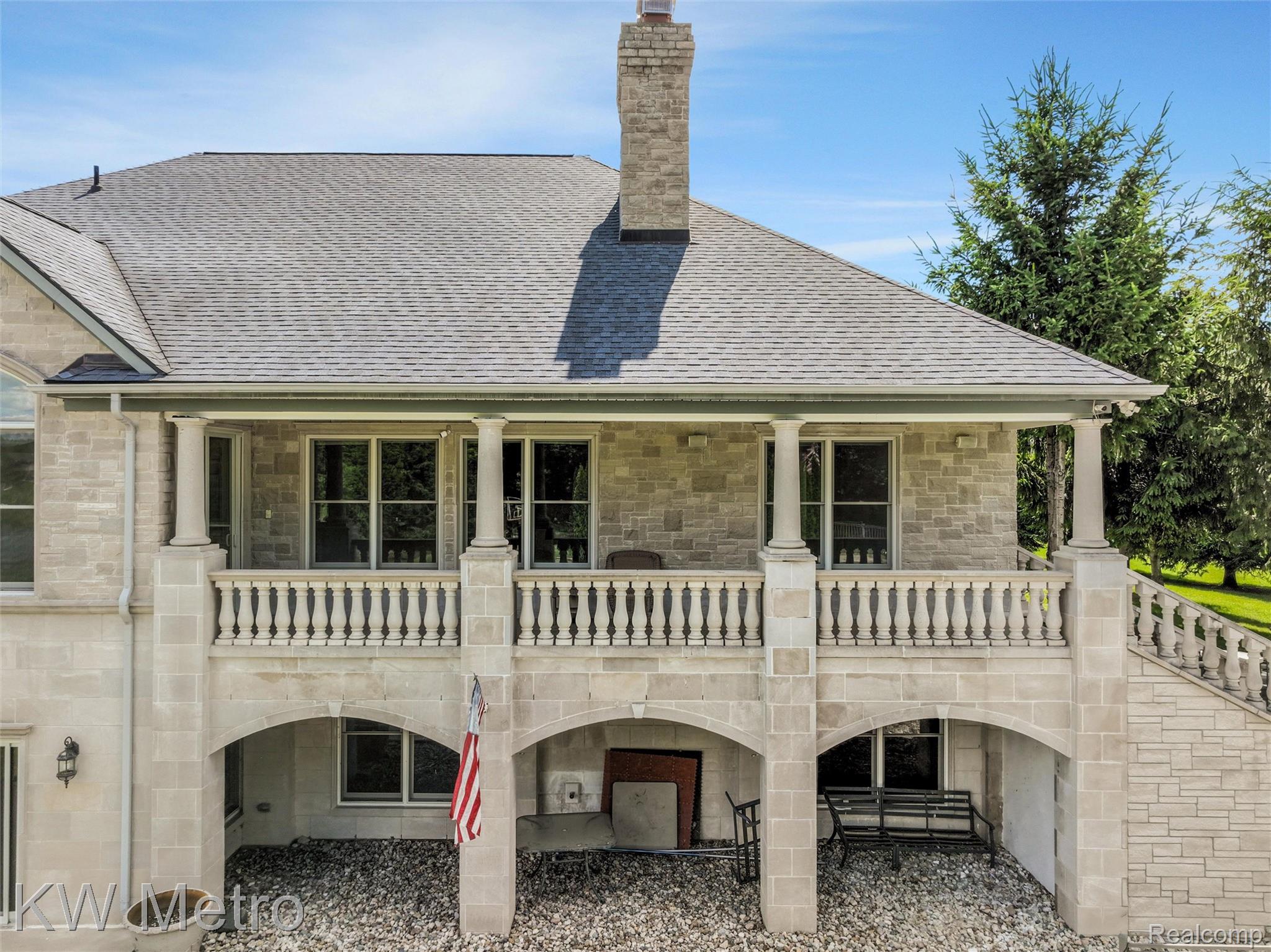 65884 Windrose Court Ray, MI 48096 - Photo 8 of 82 Stone facade residence featuring a covered second-story balcony with balustrade railing, lower-level covered patio with arched openings, and a prominent stone chimney