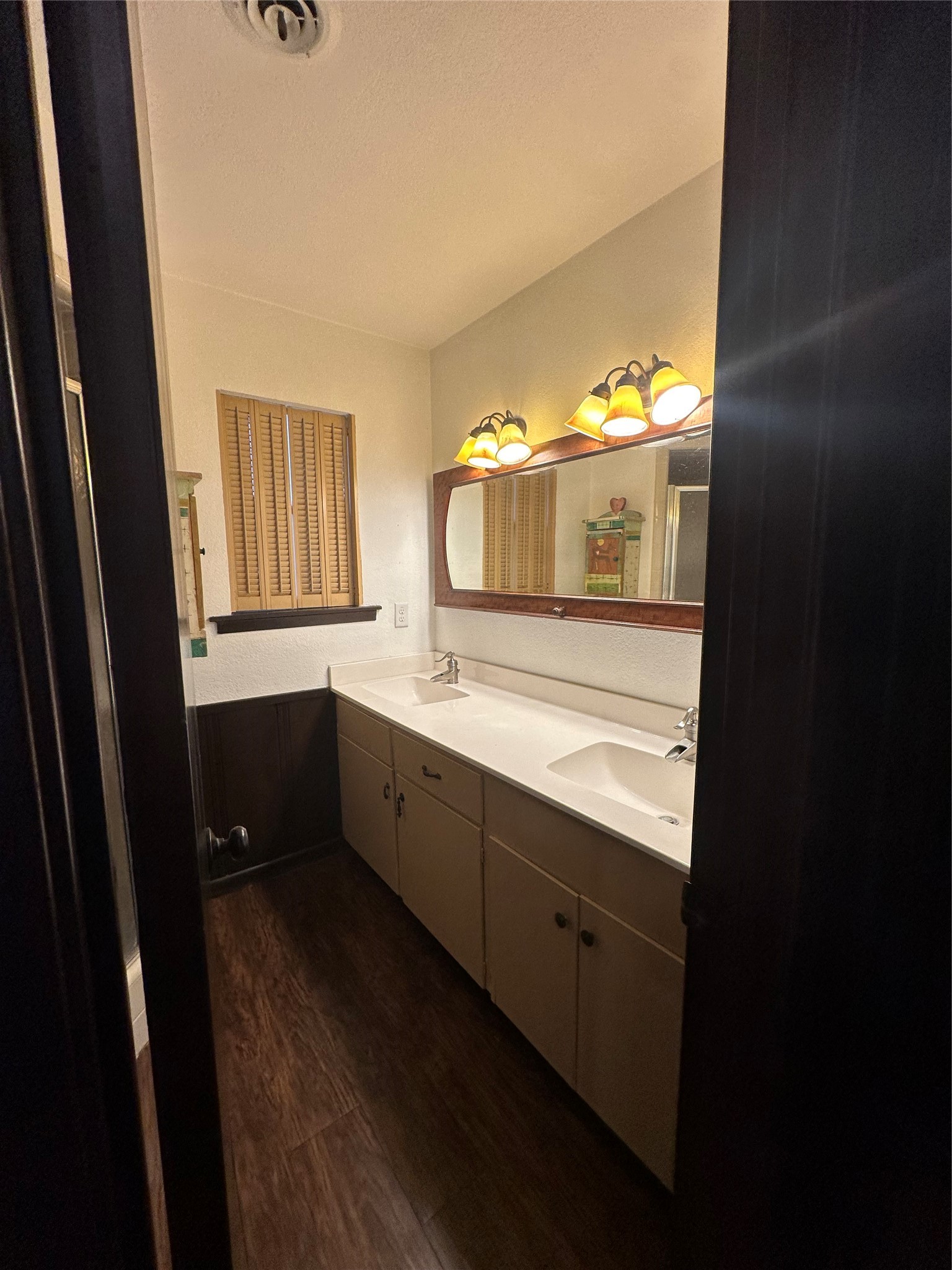 1604 Hailey Street, Unit B Conroe, TX 77301 - Photo 6 of 9 The spacious primary bathroom includes a long, beige double vanity with abundant storage cabinet doors and drawers. It is brightly lit by multi-bulb vanity lights and features plantation-style shutters over the window and a large mirror with warm wooden trim.