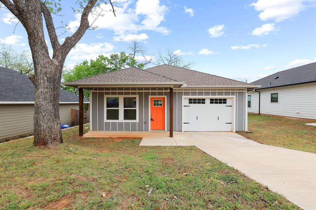 1221 West Johnson Street Denison, TX 75020 - Photo 1 of 1 Single-story residence featuring light gray siding with vertical accents, an orange front door, a single-car garage with a white door, and a concrete driveway