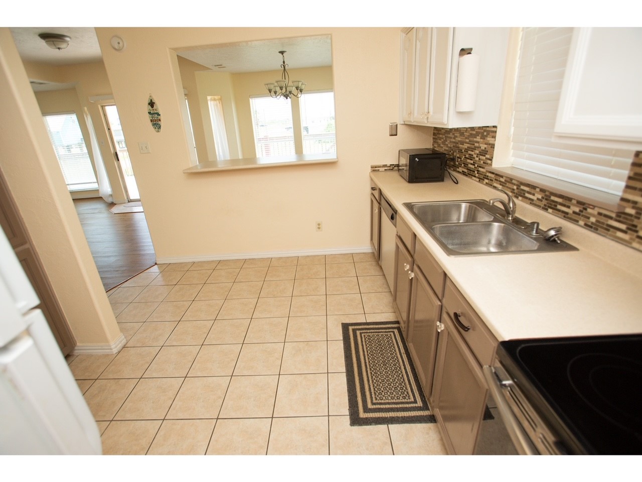 419 Thunder Road Surfside Beach, TX 77541 - Photo 13 of 32 This kitchen features a clean, open layout with beige tile flooring and neutral-colored cabinetry. It includes a double sink, modern appliances, and a pass-through window to a bright dining area, enhancing the flow and connectivity of the space.