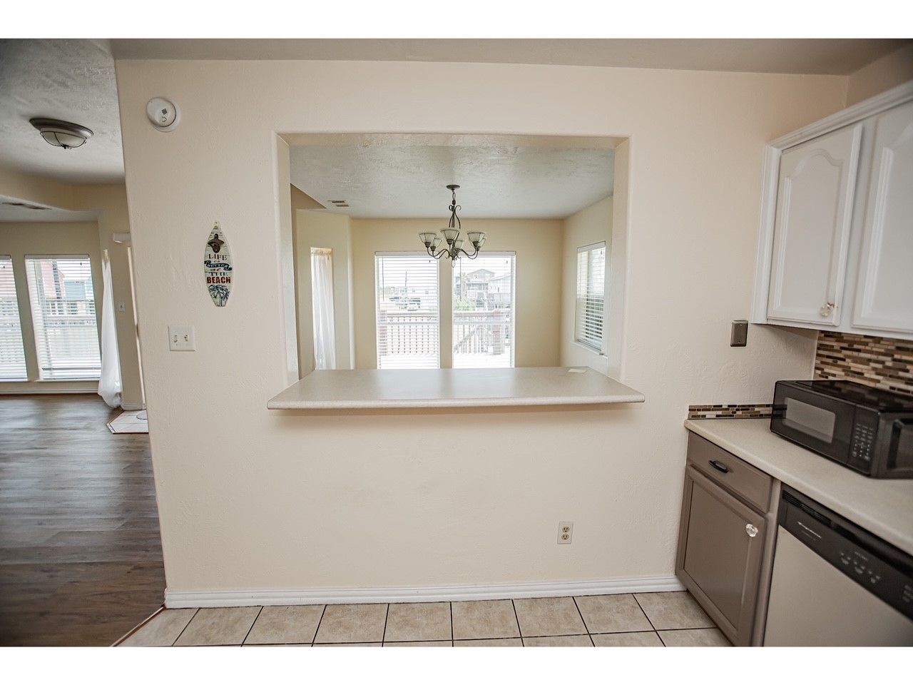 419 Thunder Road Surfside Beach, TX 77541 - Photo 14 of 32 This photo showcases a kitchen with a passthrough window to a bright dining area, featuring tile flooring and white cabinetry. The kitchen includes modern appliances and a decorative backsplash. The dining area is well-lit with large windows and a central light fixture, adding to the home's open feel.