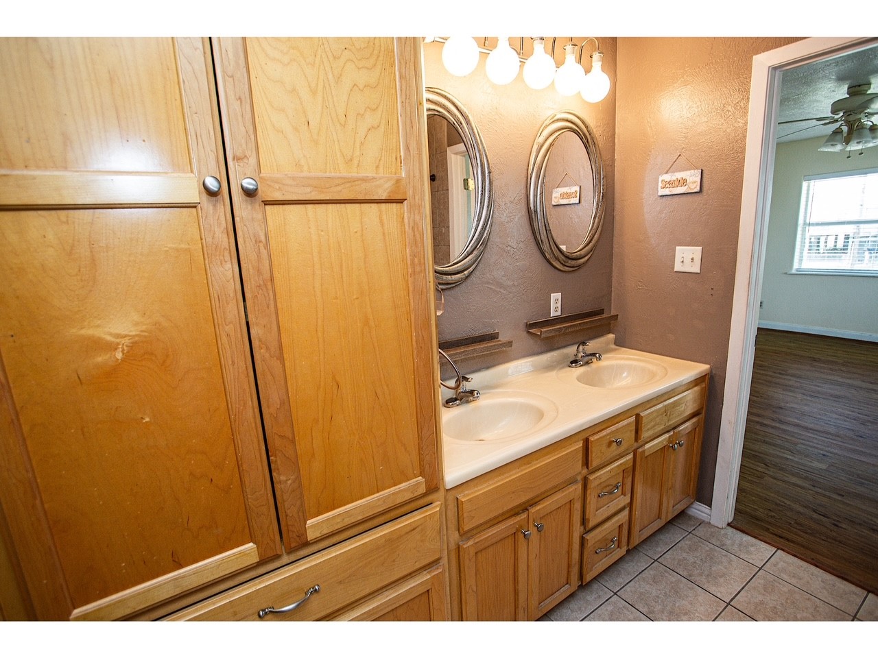 419 Thunder Road Surfside Beach, TX 77541 - Photo 18 of 32 This bathroom features dual sinks with oval mirrors, ample wooden cabinetry, and tile flooring. It opens to a room with wood flooring and a ceiling fan, providing a warm, cohesive look.