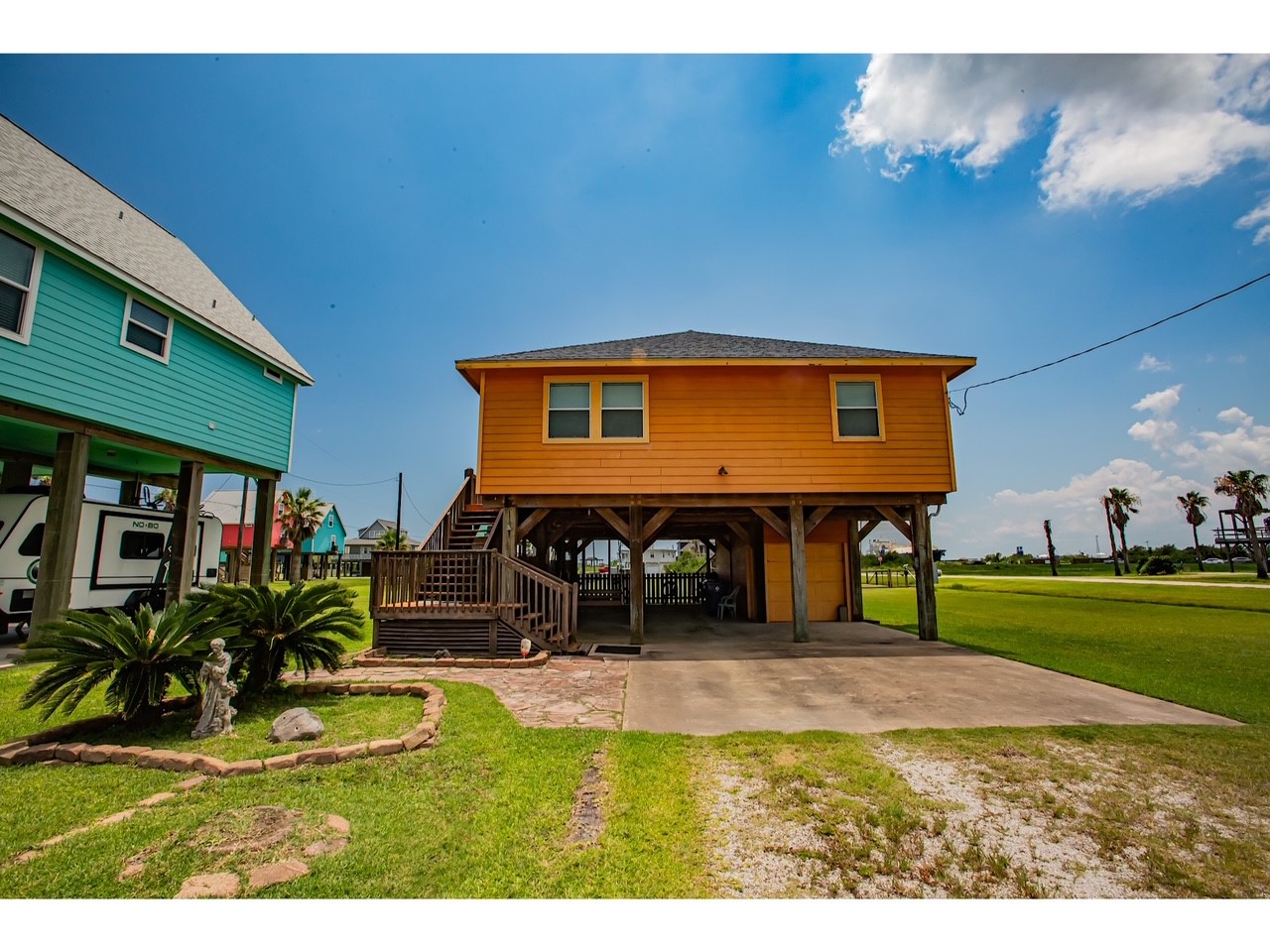 419 Thunder Road Surfside Beach, TX 77541 - Photo 25 of 32 Bright elevated home with a vibrant orange exterior, featuring a covered parking area underneath. It includes a landscaped front yard with a small garden and palm trees. The home is set in a coastal area with nearby greenery and open space.