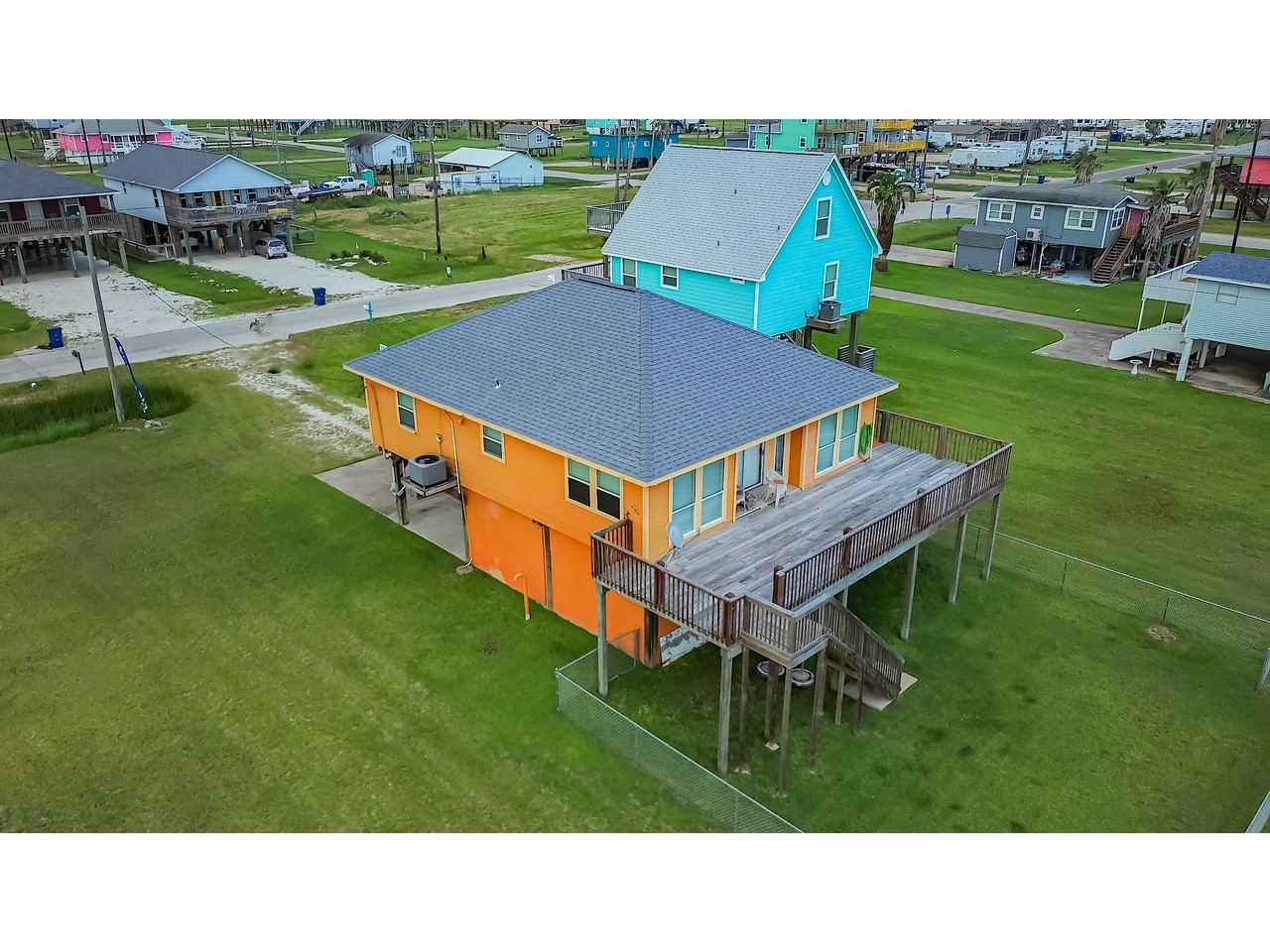 419 Thunder Road Surfside Beach, TX 77541 - Photo 26 of 32 This elevated home features a vibrant orange exterior and a spacious wooden deck, perfect for relaxation or entertaining. It's situated in a neighborhood with other colorful homes, offering a charming and lively setting. The surrounding area is grassy, providing ample outdoor space.