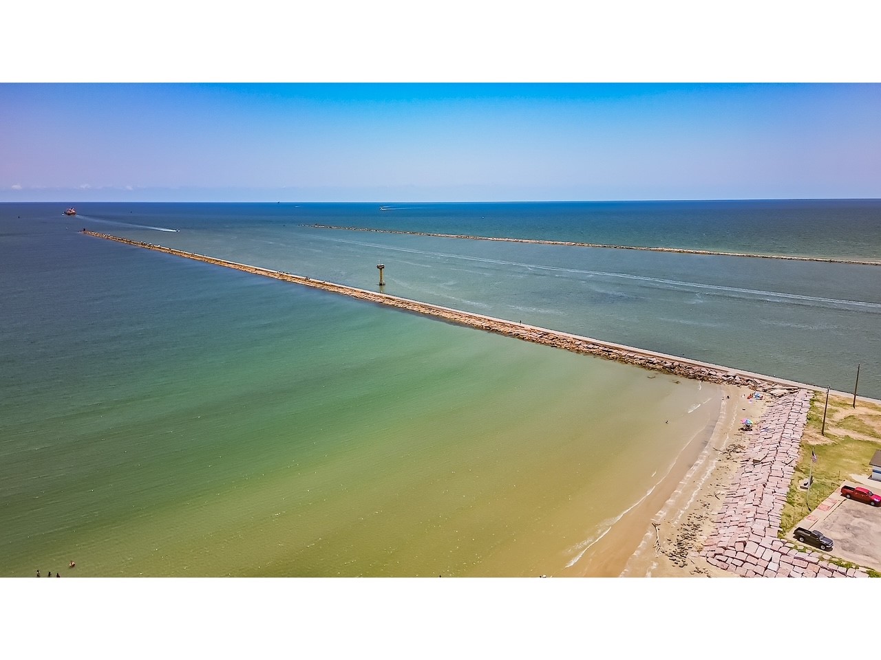 419 Thunder Road Surfside Beach, TX 77541 - Photo 32 of 32 Aerial view of a coastal area with a long jetty extending into the ocean, bordered by clear blue and green waters. The shoreline features a sandy beach with a few parked cars, offering a serene and scenic waterfront setting.