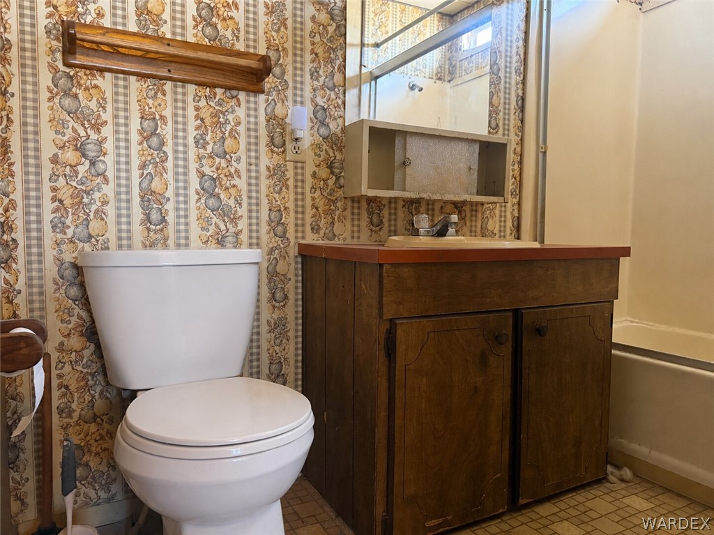 1666 Talc Road Bullhead City, AZ 86442 - Photo 31 of 61 Bathroom featuring a white toilet, wood-finish vanity with a red-toned countertop, wall-mounted mirror, and patterned wallpaper
