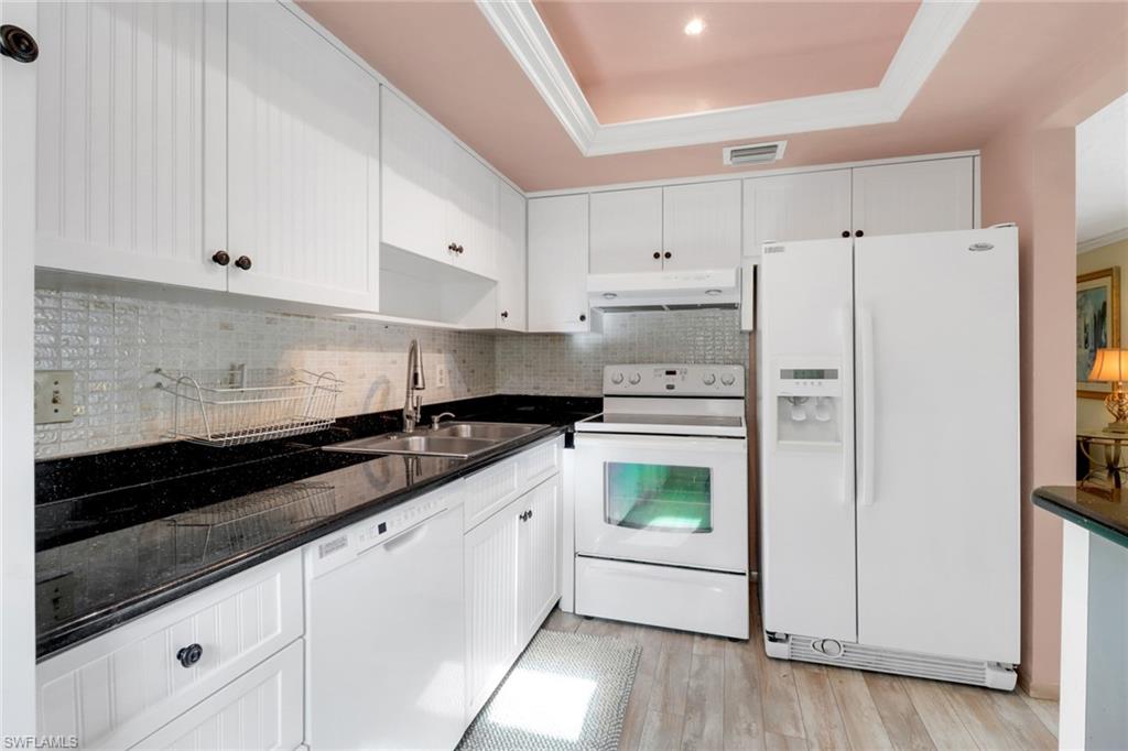 1155 Sandpiper Street, Unit D4 Naples, FL 34102 - Photo 7 of 39 Kitchen with white appliances, under cabinet range hood, a tray ceiling, dark countertops, and decorative backsplash