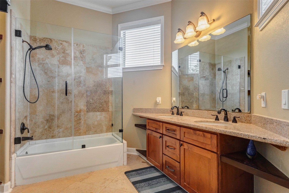 5610 Westerham Weston Lakes, TX 77441 - Photo 31 of 47 The ensuite bath features a granite countertop vanity with dual sinks, wood cabinetry, and a tub and shower combination with glass enclosure and tile surround. A window floods the space with natural light.