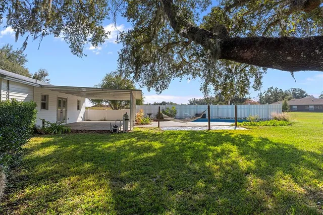 a view of a white house in front of a big yard with plants and large trees